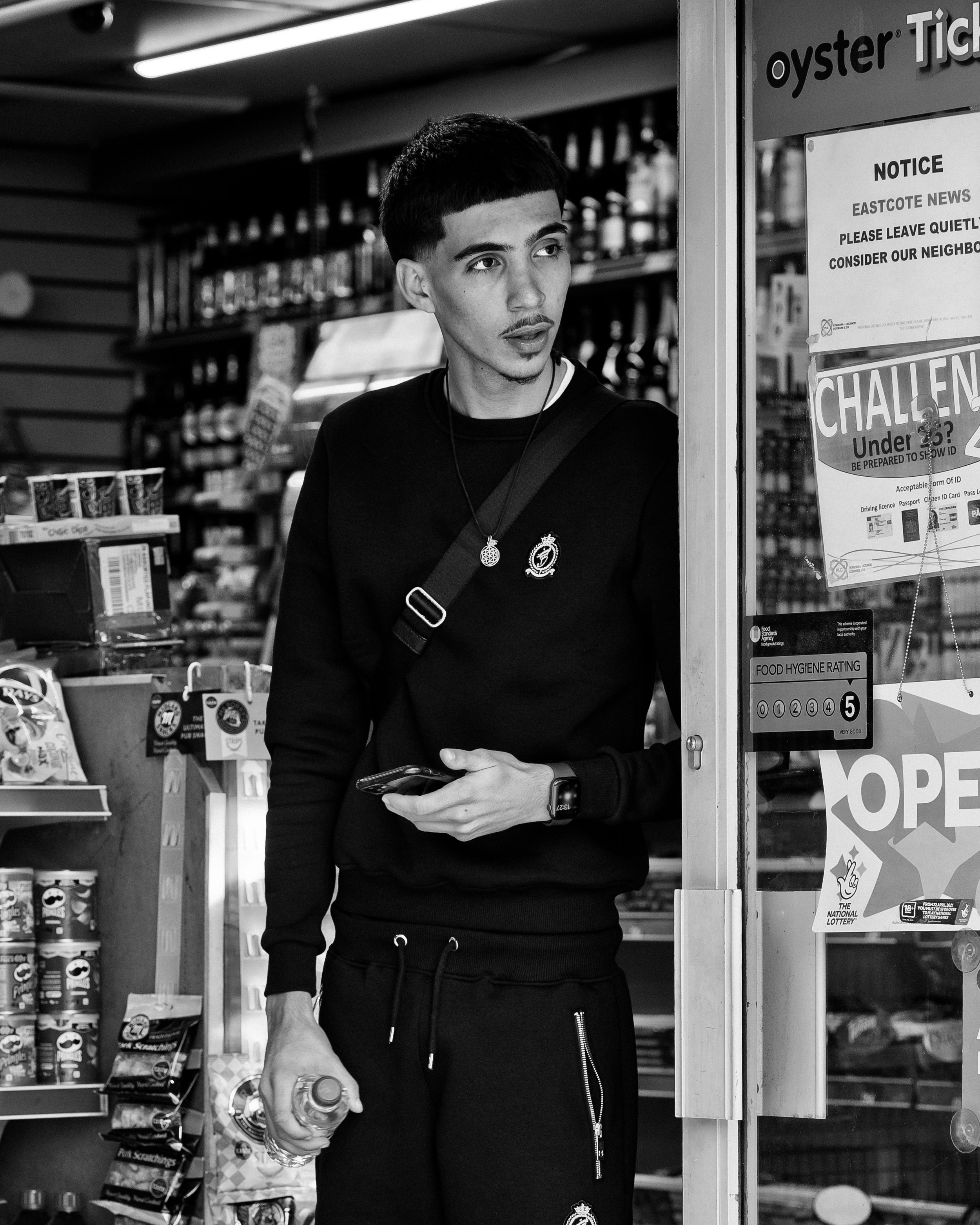 A young man standing in a convenience store holding a bottle of water in his left hand and a phone in his right hand, looking towards the right. He is dressed in a black sweatshirt with a logo, black sweatpants, and has a shoulder bag.