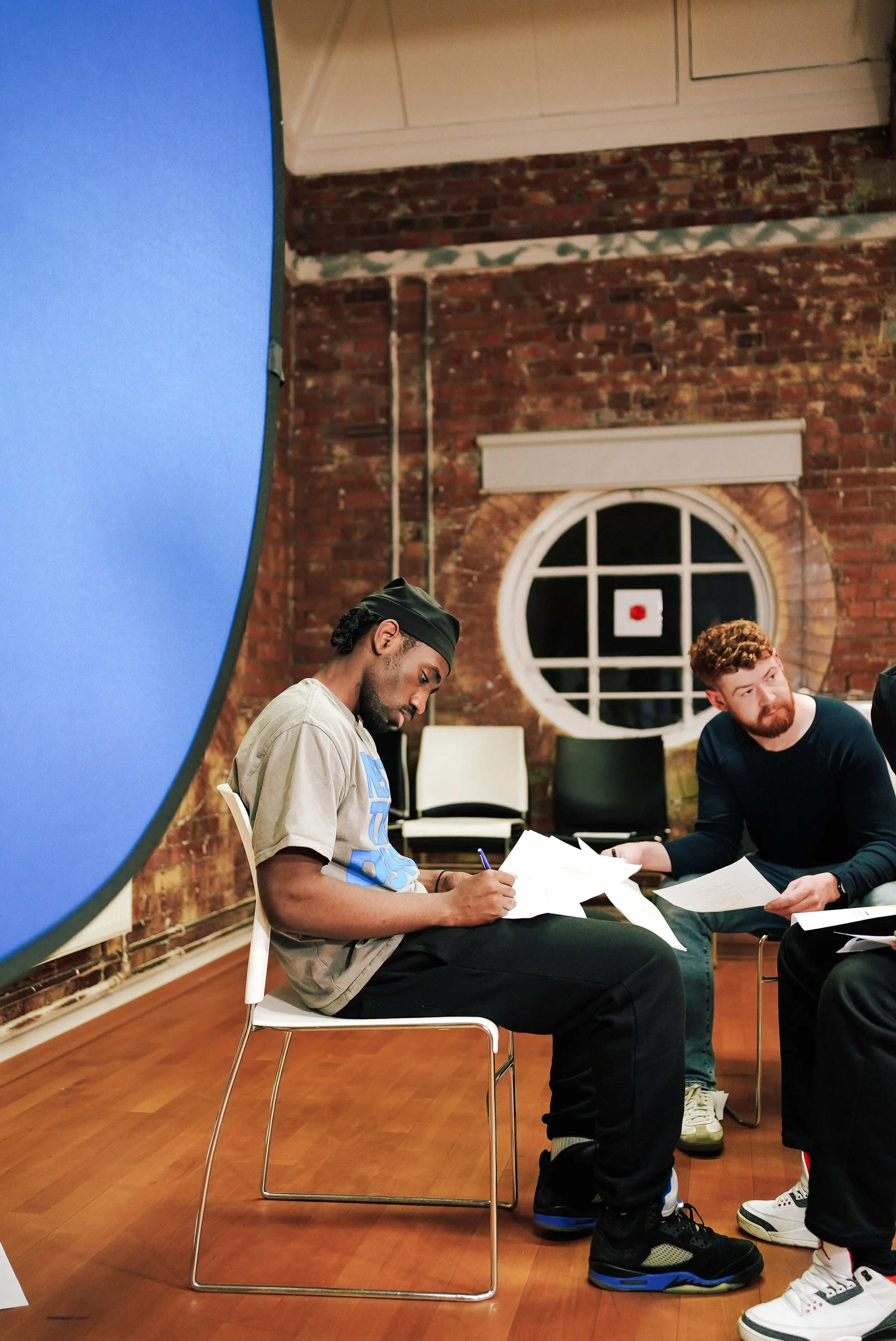 Two men sitting on chairs during a rehearsal or practice, reviewing papers, in a room with brick walls and a large circular window.