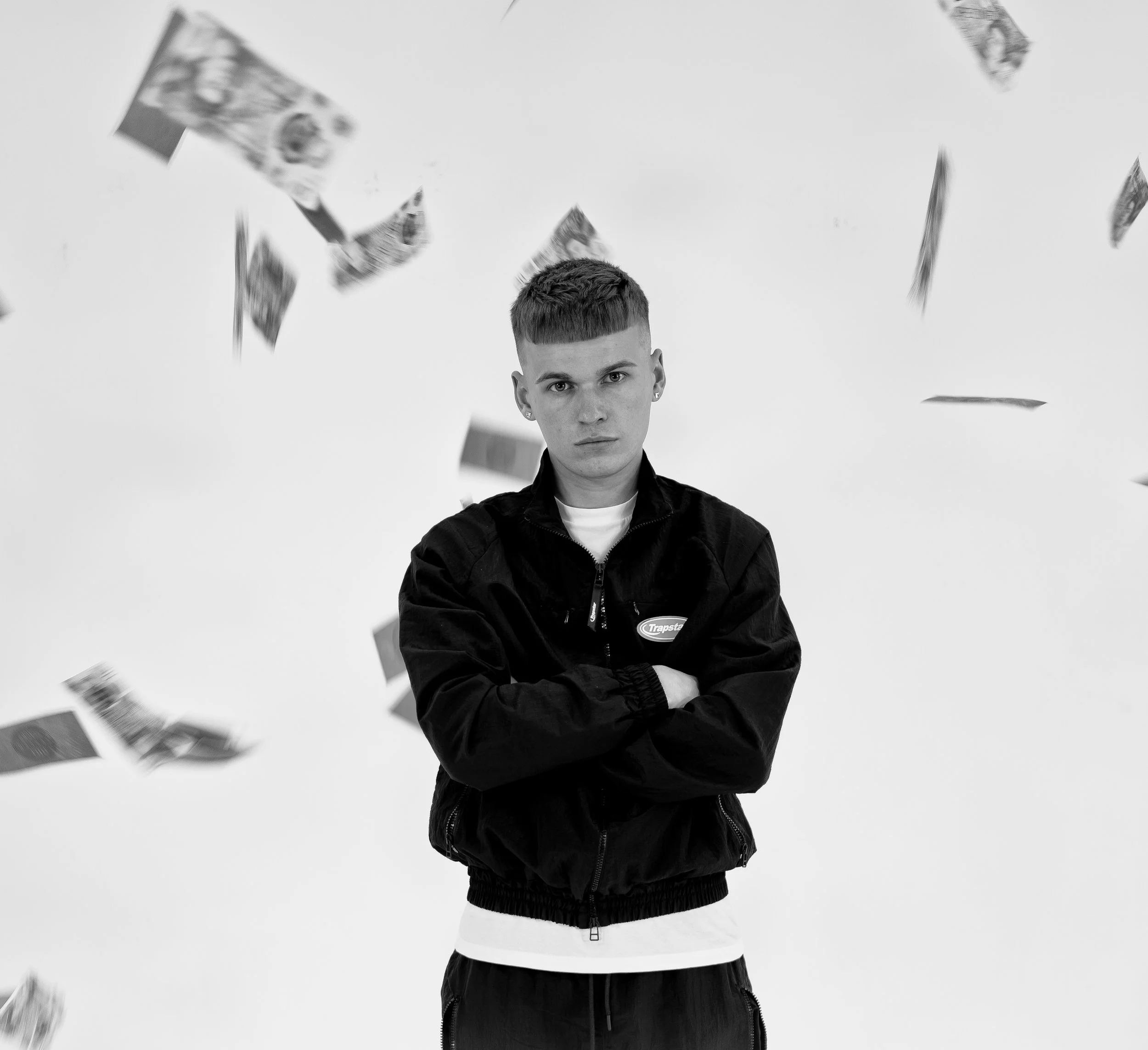 Black and white photo of a young man with short hair, wearing a black jacket with arms crossed, standing amidst falling dollar bills against a plain background.