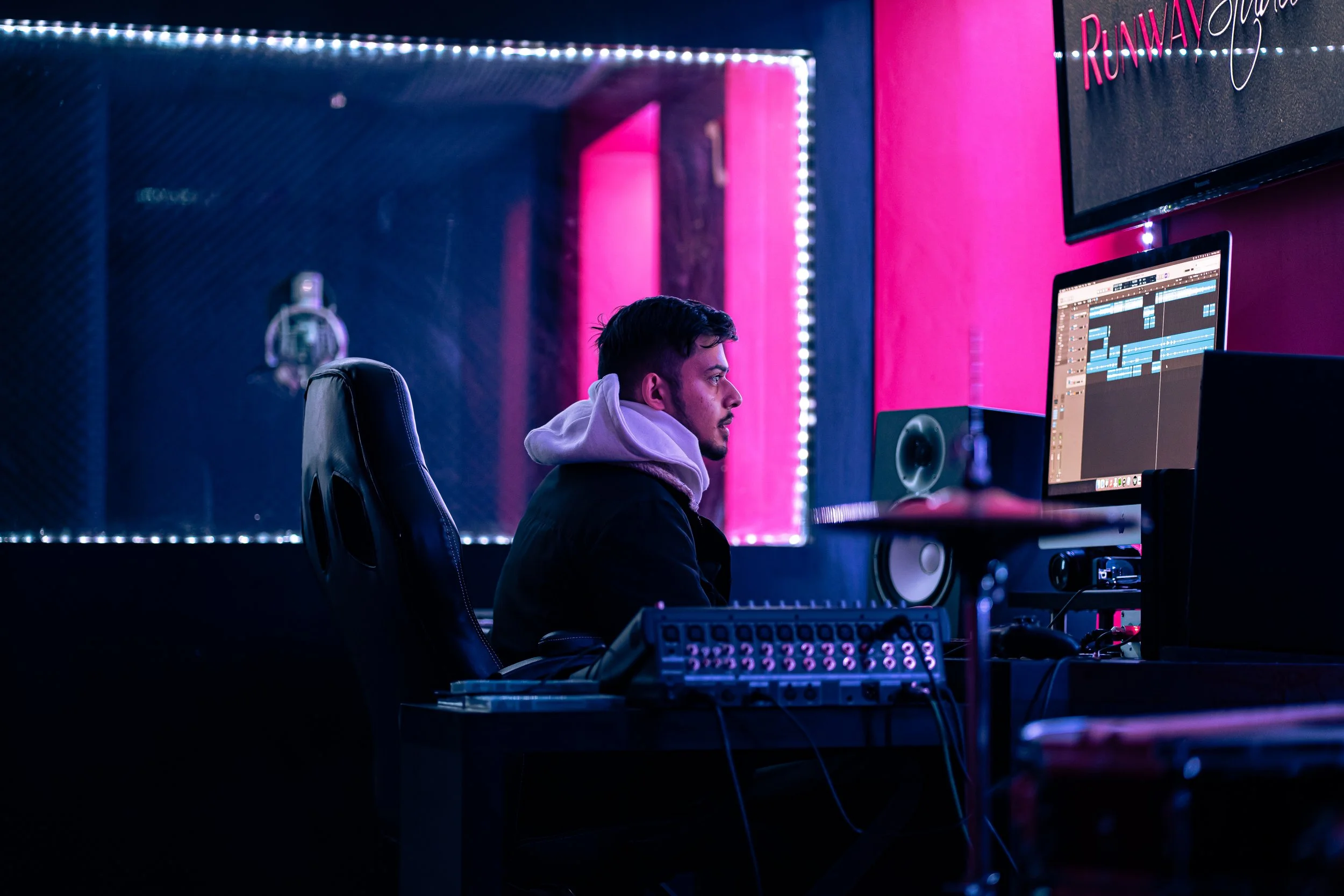 A young man working on music production in a darker, neon-lit recording studio with pink and purple lighting, focused on a computer screen displaying audio editing software.