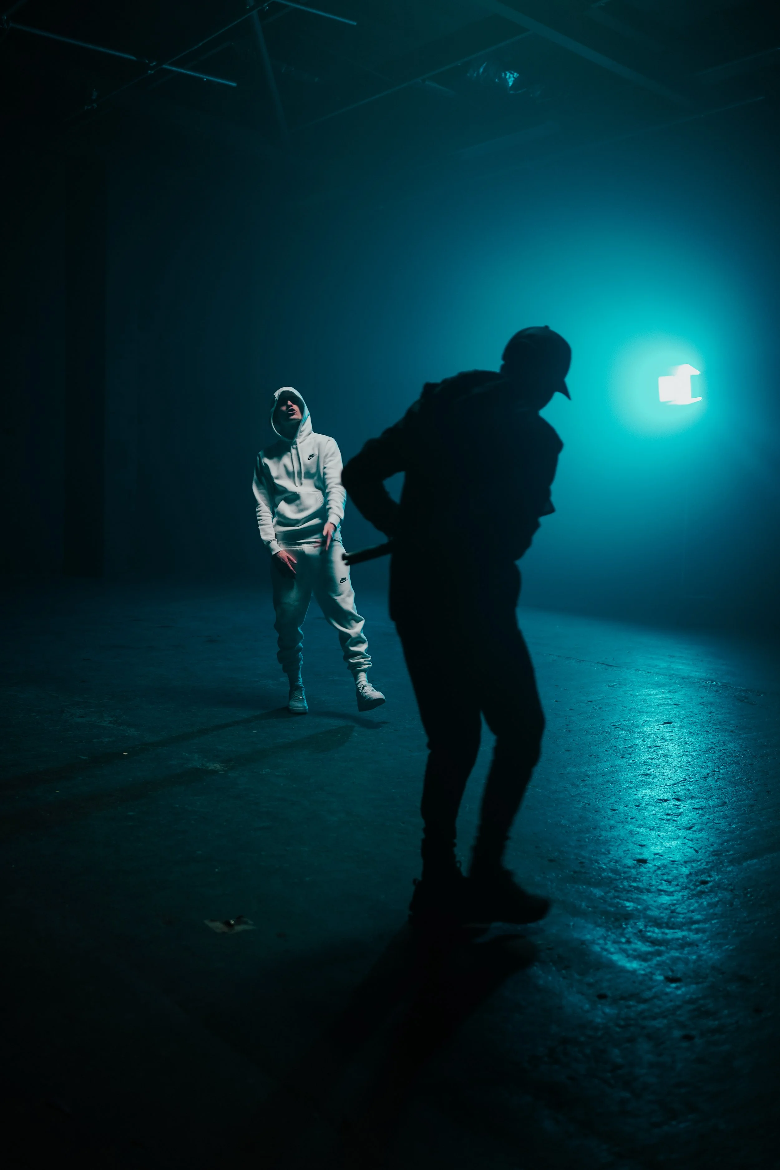 A person in a white hoodie and sweatpants stands in the background, while another person wearing dark clothing and a cap, holding an object, appears to be walking in the foreground. The scene is dark with blue lighting.