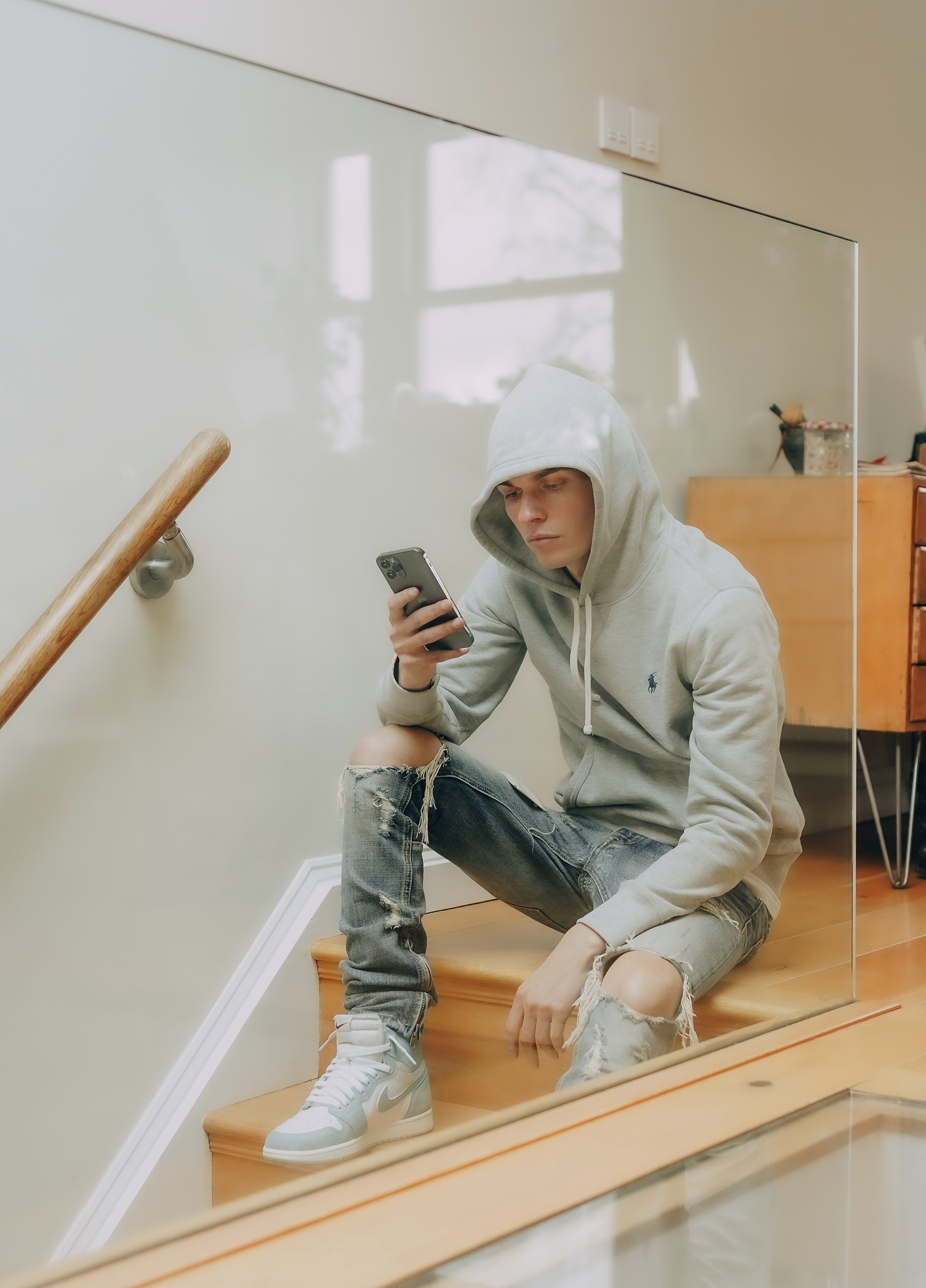 Young man sitting on wooden stairs, wearing a gray hoodie with the hood up, ripped jeans, and Nike sneakers, looking at a smartphone.