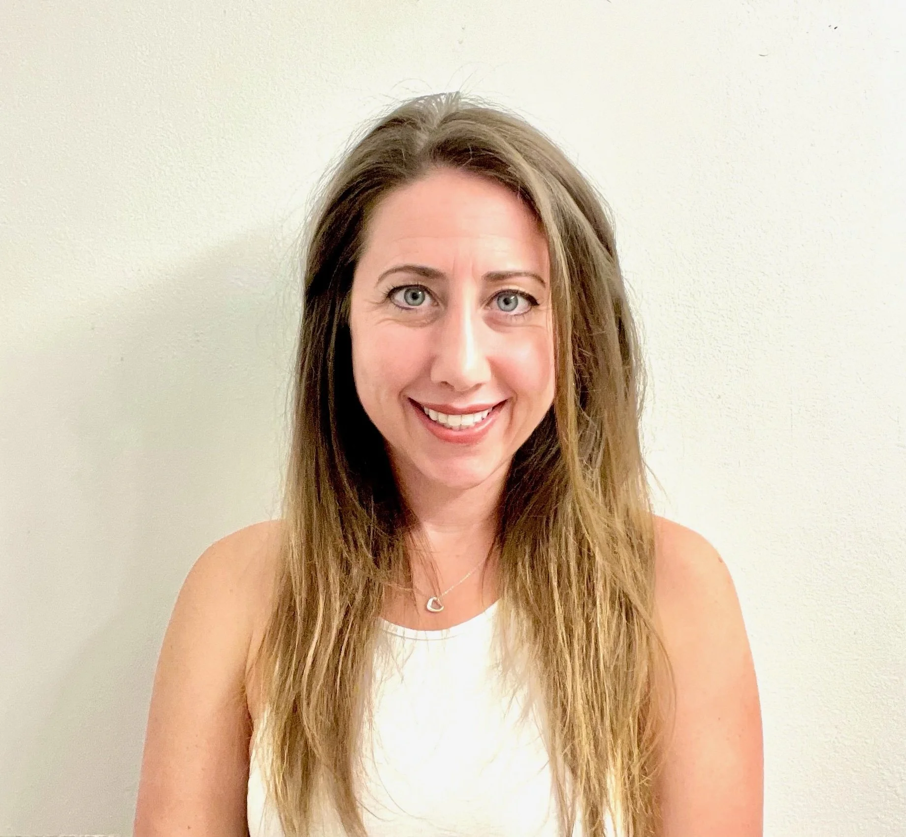 A woman with long, wavy brown hair and blue eyes smiling at the camera against a plain cream-colored wall.