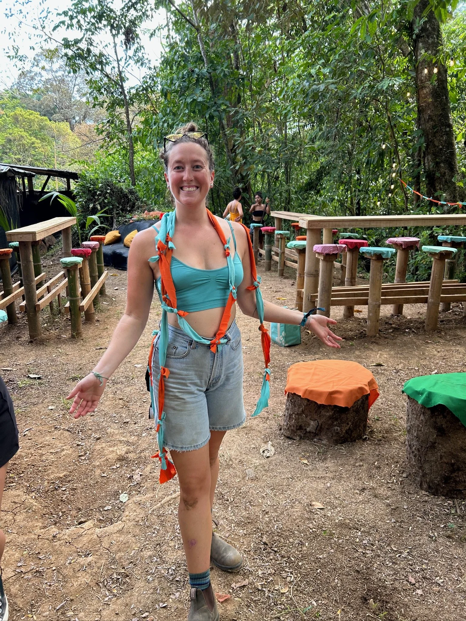 Smiling woman in teal crop top and denim shorts standing outdoors in a forested area with wooden benches and colorful painted stumps.