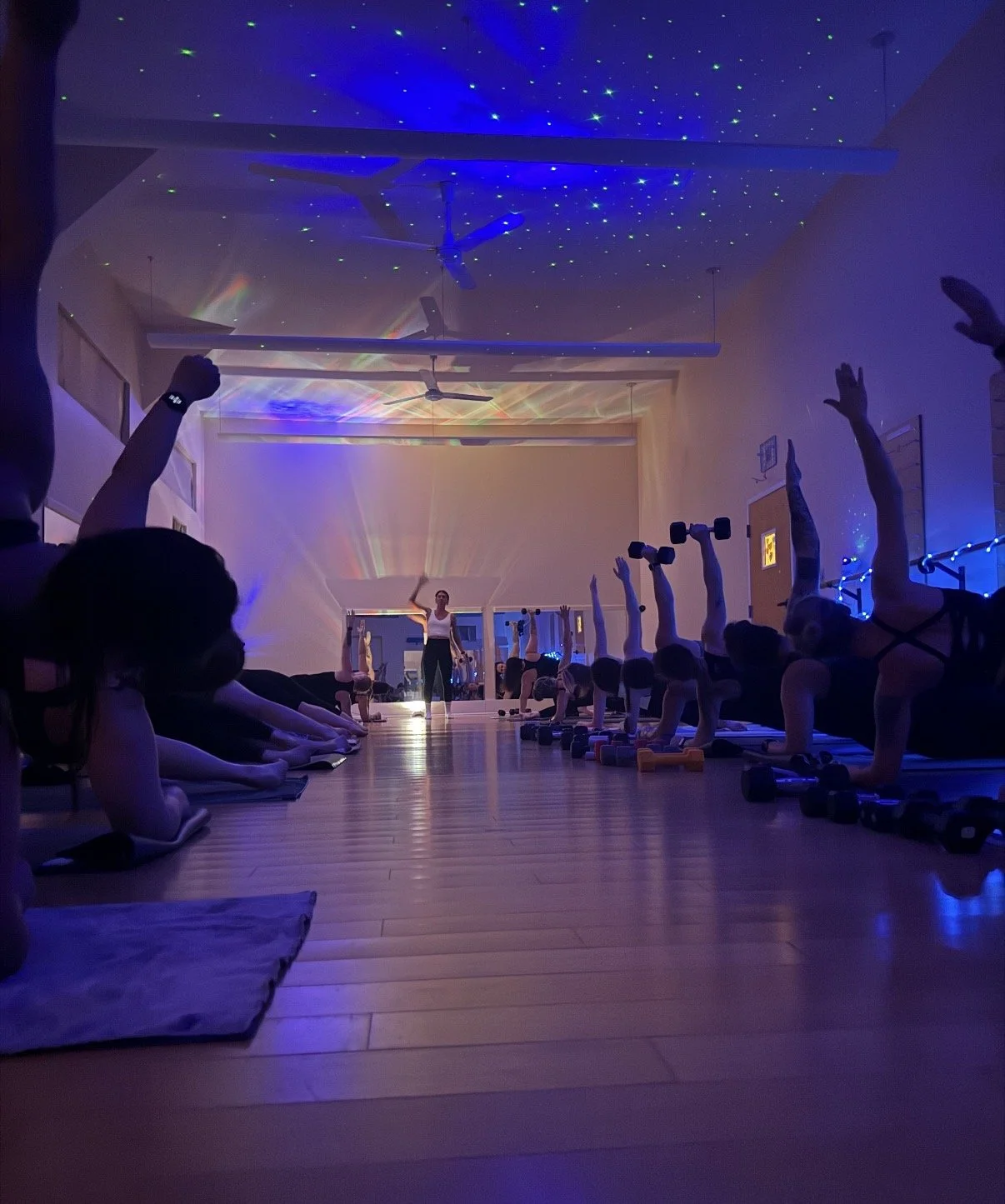 A group fitness class at night with students doing plank poses and lifting weights in a dark room decorated with starry lights and a rainbow projection.