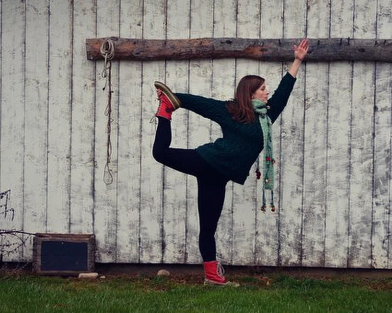 A woman practicing yoga outdoors in front of a weathered wooden wall, holding her leg behind her and reaching up with her arm.
