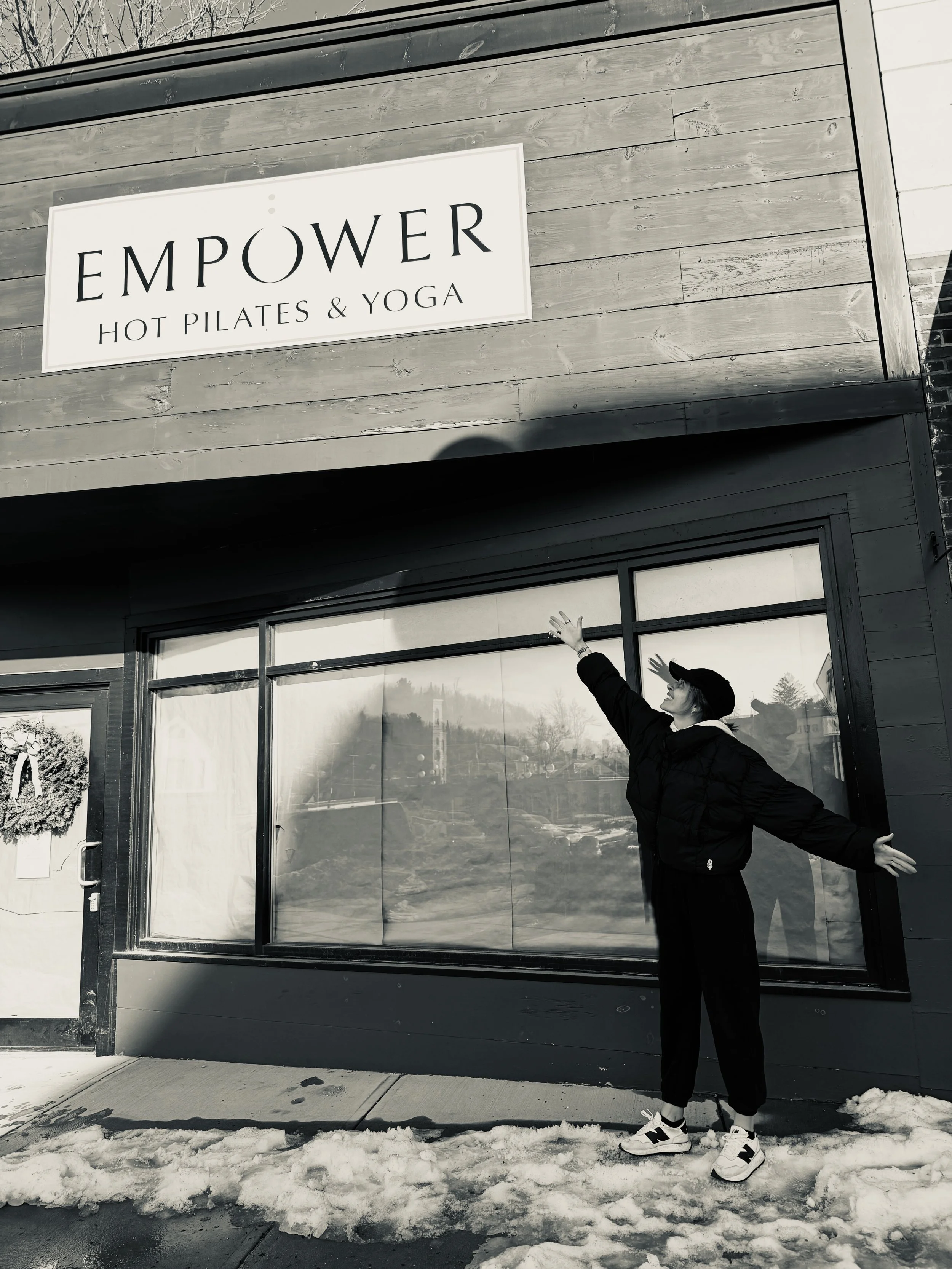 A woman standing outside a building with a sign reading "EMPOWER HOT PILATES & YOGA," posing with arms outstretched in front of a window, with snow on the ground.