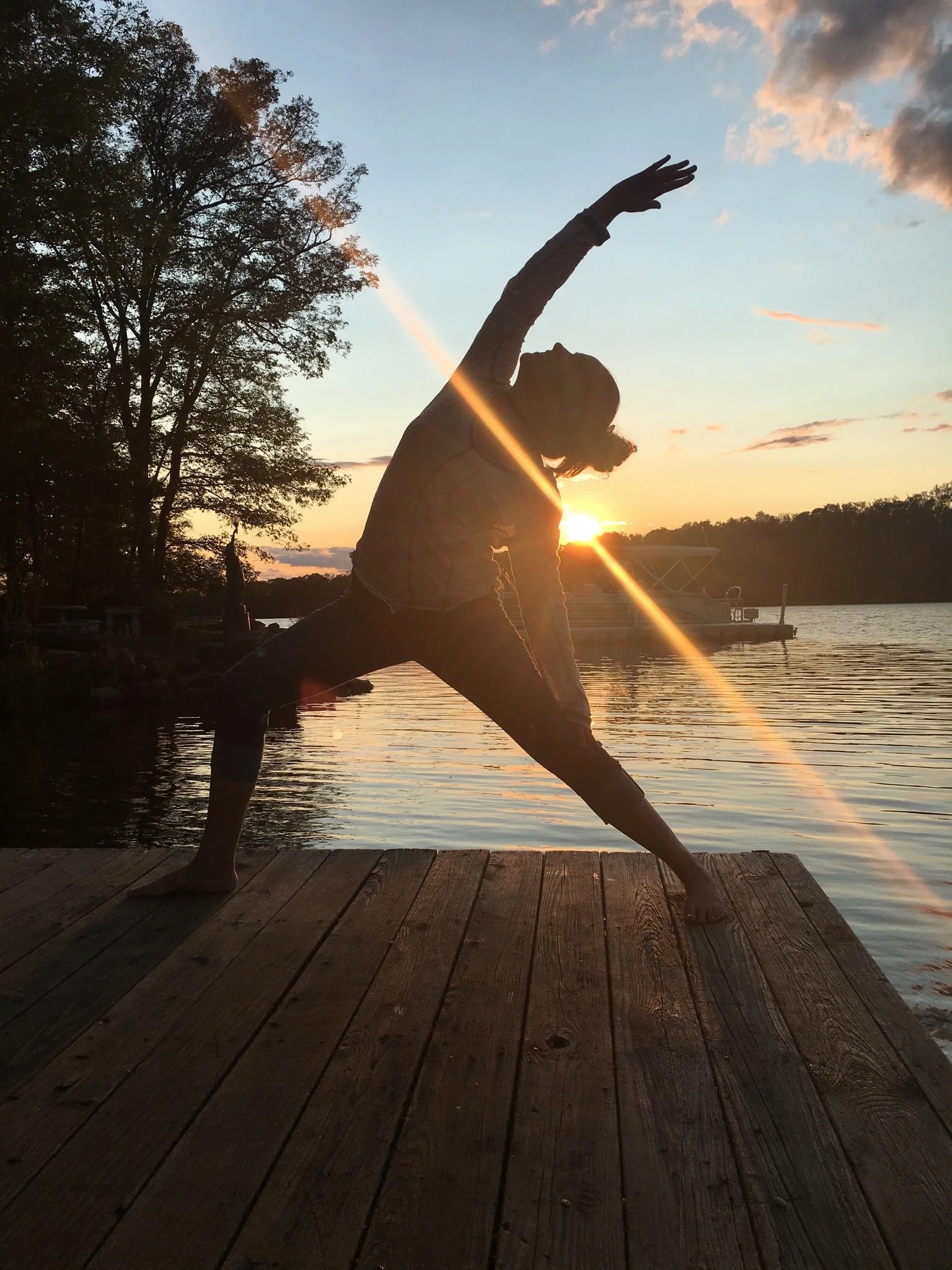 Person practicing yoga on a wooden dock at sunset by a lake, with trees and a boat in the background.