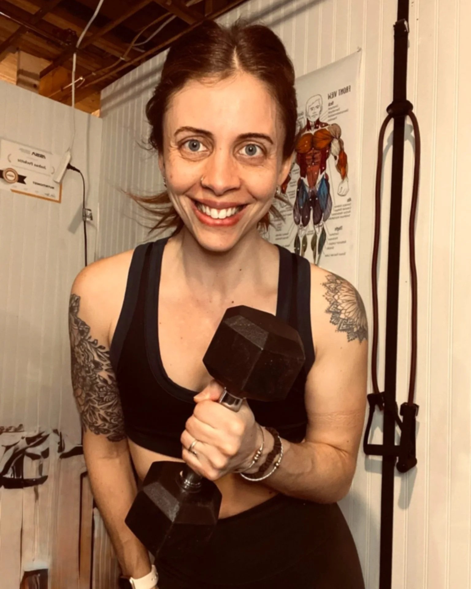 A woman with short reddish hair, smiling, holding a black dumbbell, standing in a gym with a anatomical poster and workout straps in the background.