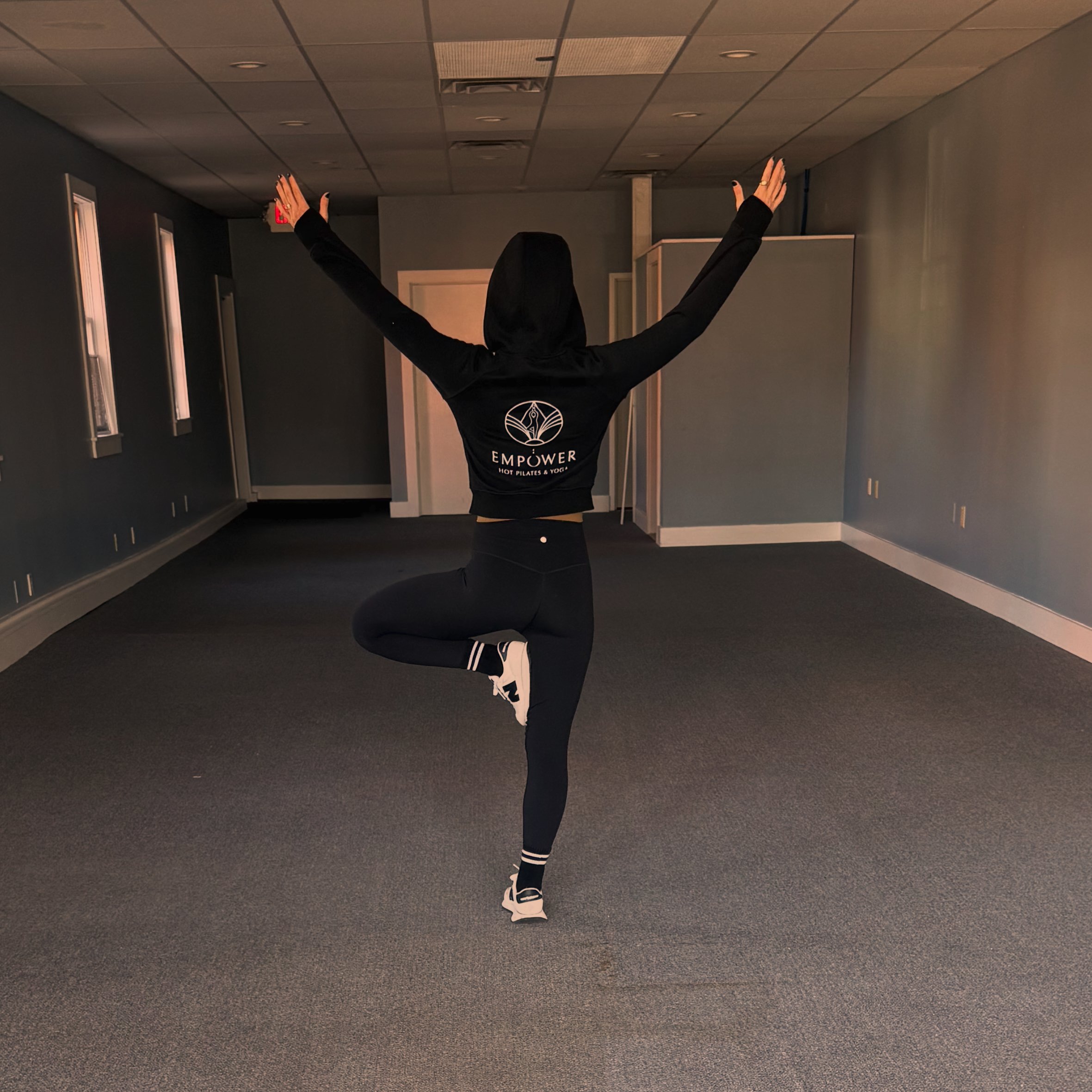 Person standing on one leg in a yoga pose in an empty room, wearing black leggings, a black hoodie with a logo, and sneakers, with arms raised and outstretched.