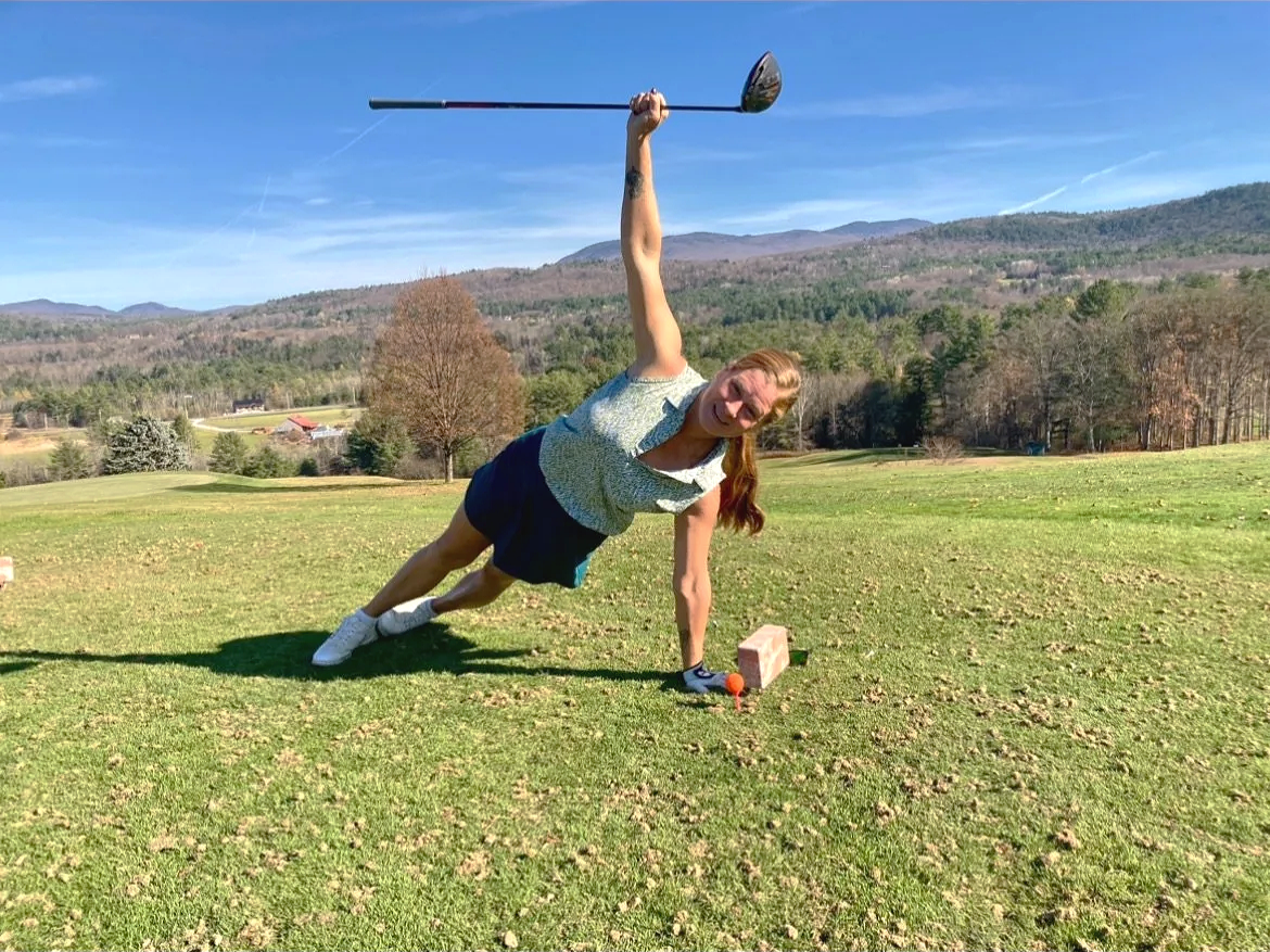 Woman performing a side plank exercise outdoors on a grassy field, holding a golf club in one hand with mountains and trees in the background.