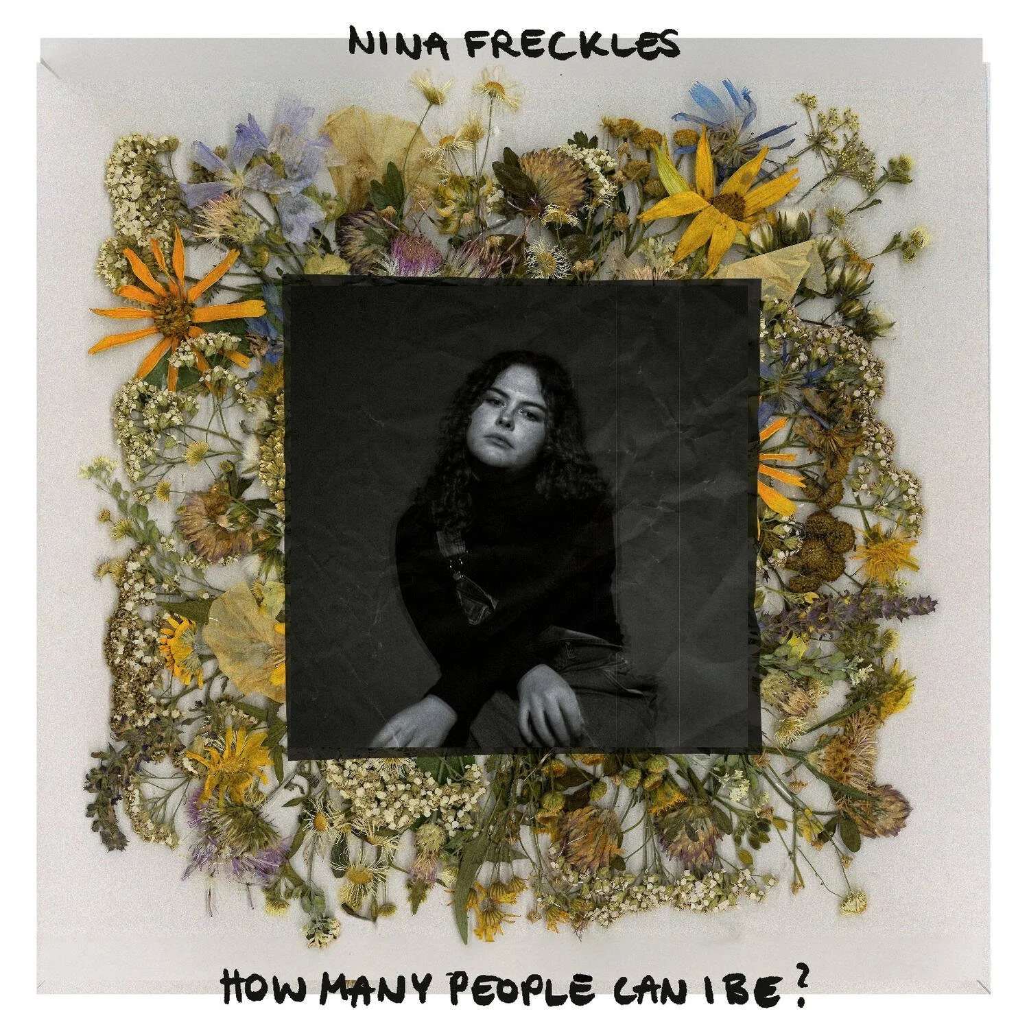 Schwarzes und weißes Portrait eines jungen Mädchens mit lockigem Haar, das auf einer welligen Oberfläche sitzt, vor einem Hintergrund aus einer Vielzahl von getrockneten Blumen. Oben steht in schwarzer Handschrift 'Nina Freckles', unten in Großbuchstaben 'How many people can be?'