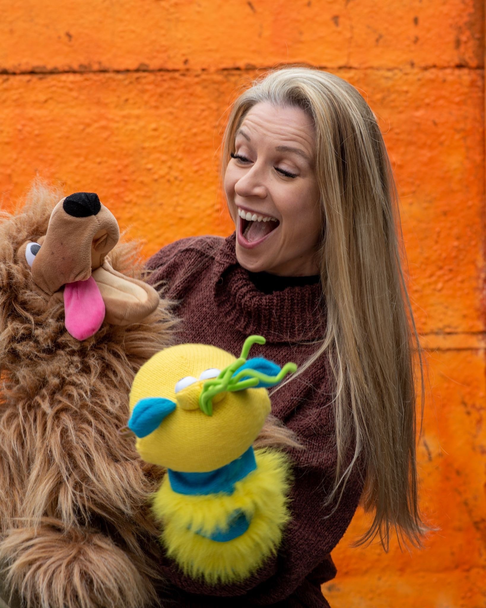 A woman with long, blonde hair smiling as she interacts with two puppets, one resembling a lion and the other a worm, against an orange wall background.