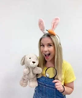 Young woman with bunny ears headband holding a plush teddy bear and a magnifying glass, smiling at the camera.