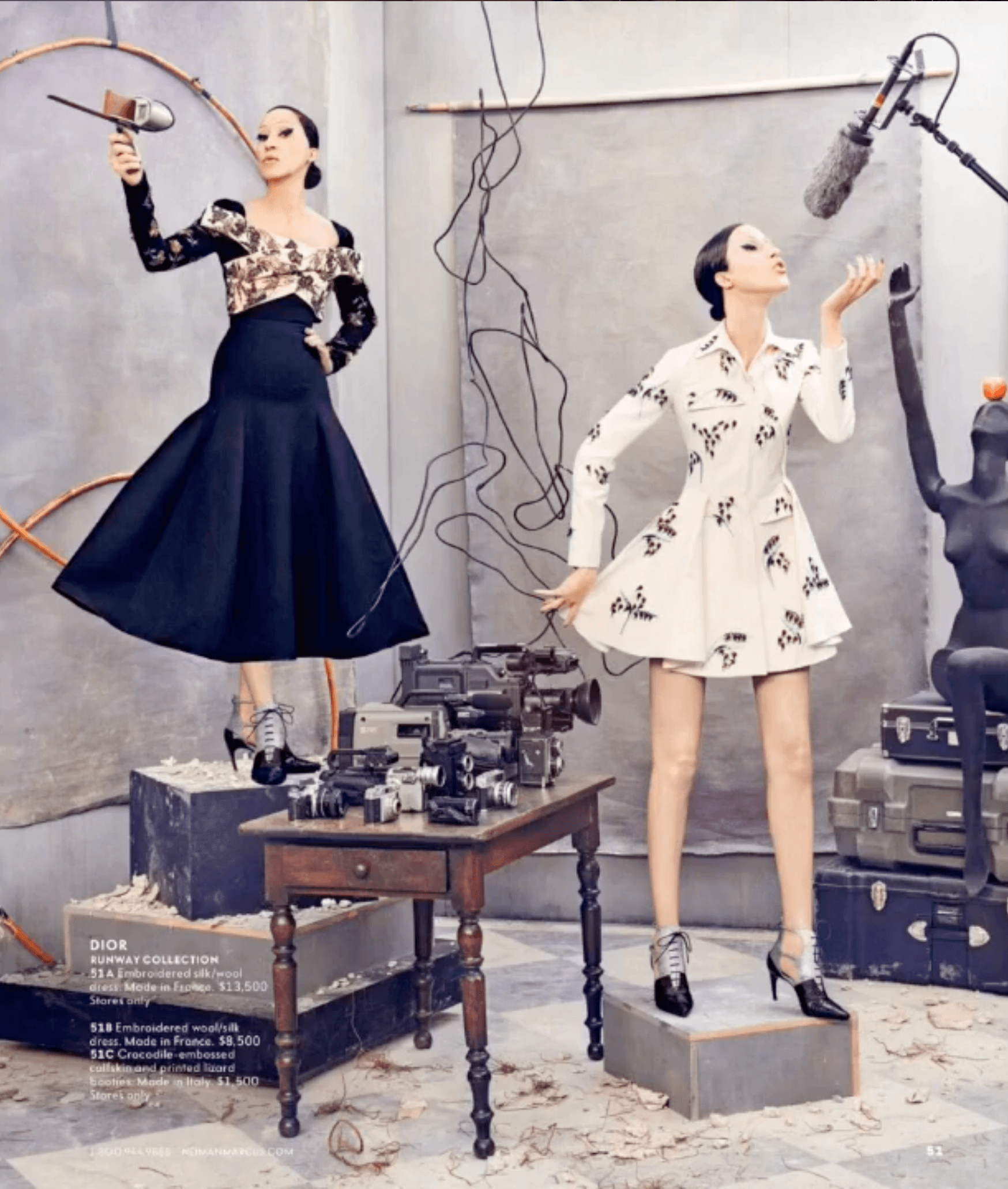 Two women in high fashion outfits are surrounded by movie production equipment. One woman stands on a platform holding a vintage microphone, while the other poses on a small block. There is a table with cameras and equipment between them, and various