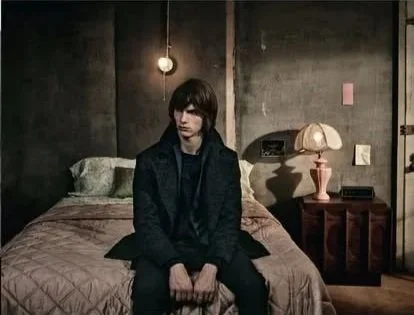 A woman with dark hair sits on the edge of a bed in a dimly lit room with dark walls. There is a lightbulb hanging from the ceiling and a nightstand with a pink lamp next to the bed.
