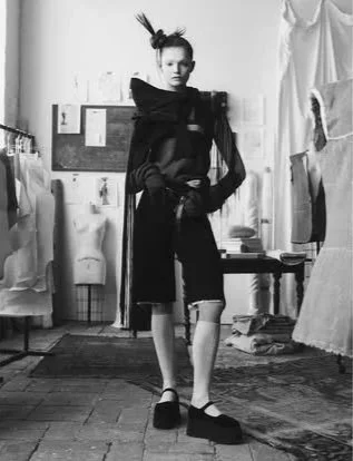 A person standing in a room with artistic and experimental fashion, wearing dark layered clothing, with their hair styled in an updo, posing in front of various furnishings and artwork.