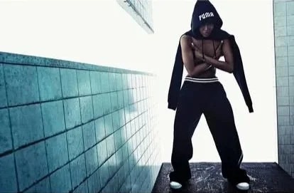 A person wearing a black hoodie, black pants, and sneakers, standing against a tiled wall and floor, with the hood covering their head and arms crossed.