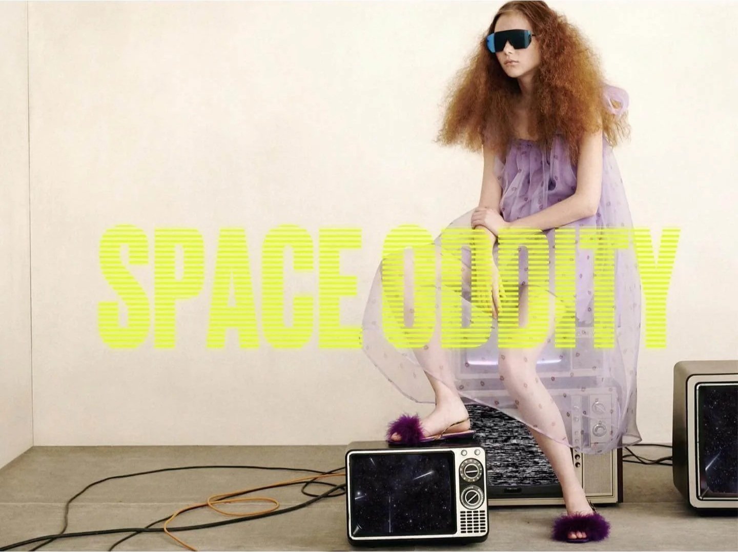 A woman with curly red hair wearing oversized sunglasses, a purple dress, and fuzzy purple slippers, sitting on vintage electronic equipment in a minimalist room with the word "SPACEODDITY" in large yellow text across the image.