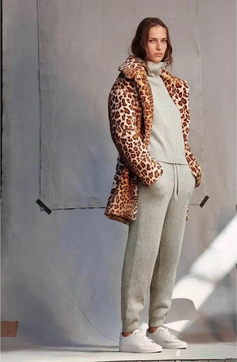 A woman standing with hands in her pockets, wearing a leopard print coat, a light gray turtleneck and matching jogger pants, and white sneakers. She has shoulder-length brown hair and a serious expression, with a plain background and fabric backdrop 