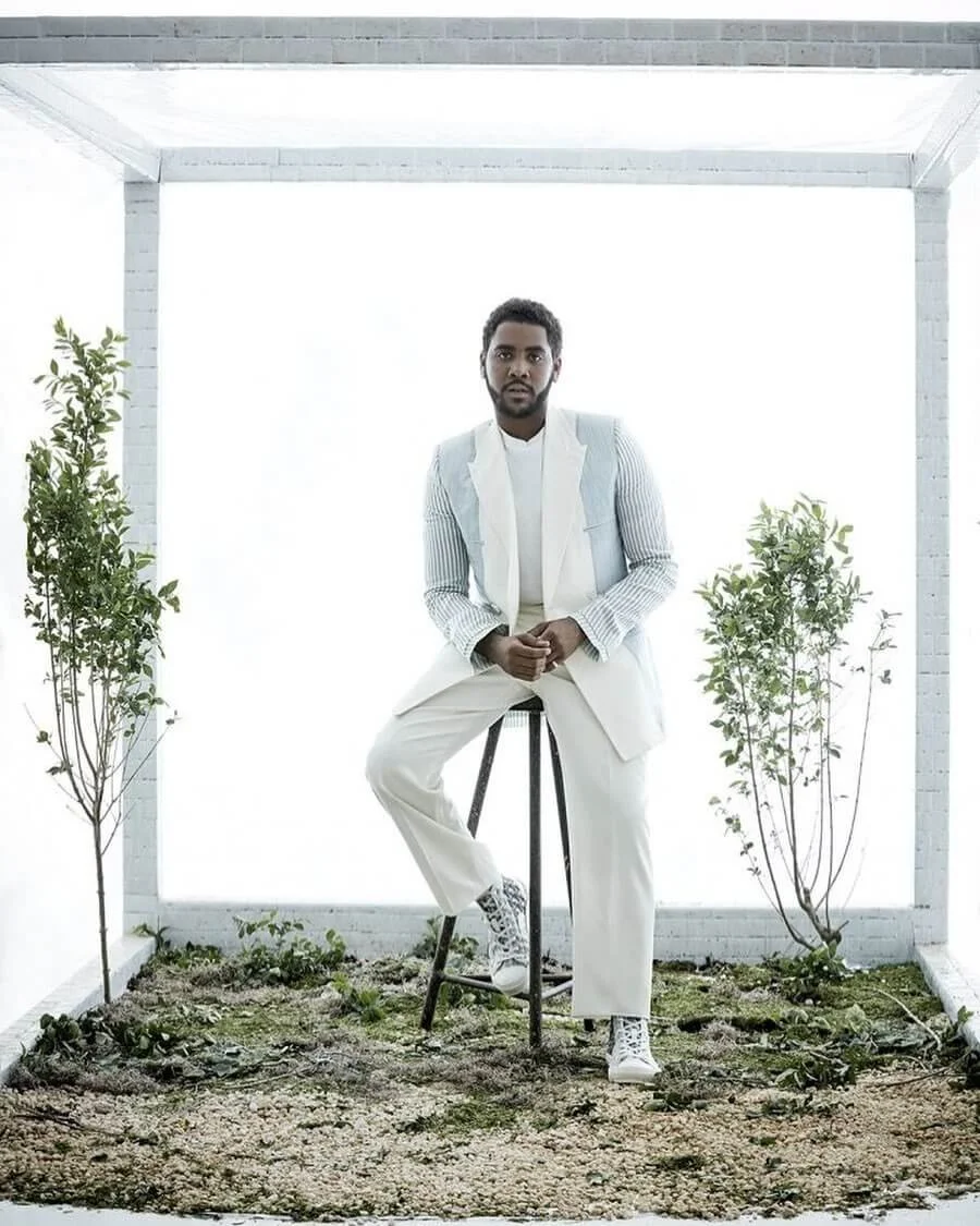 A man dressed in white sitting on a stool in a minimalistic, white indoor garden space with two small trees and a soil bed, bright lighting.