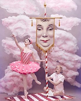 A theatrical scene with a ballerina in a pink tutu, a man holding a large portrait of a woman's face on a cloud backdrop, and pink clouds surrounding them.