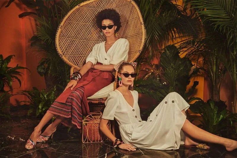 Two women posing indoors with tropical plants. One woman is seated on a wicker chair, wearing a white blouse, brown skirt, sunglasses, and jewelry. The other woman is sitting on the floor, wearing a cream dress, sunglasses, and earrings. The backgrou