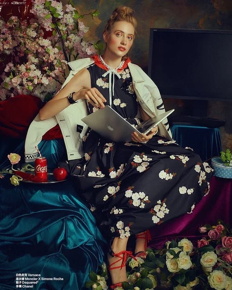 A woman wearing a black floral dress and red lace-up shoes sitting on a colorful velvet sofa, holding a silver laptop, with a floral background, a large TV, and items like a Coca-Cola can, an apple, flowers, and a plant around her.