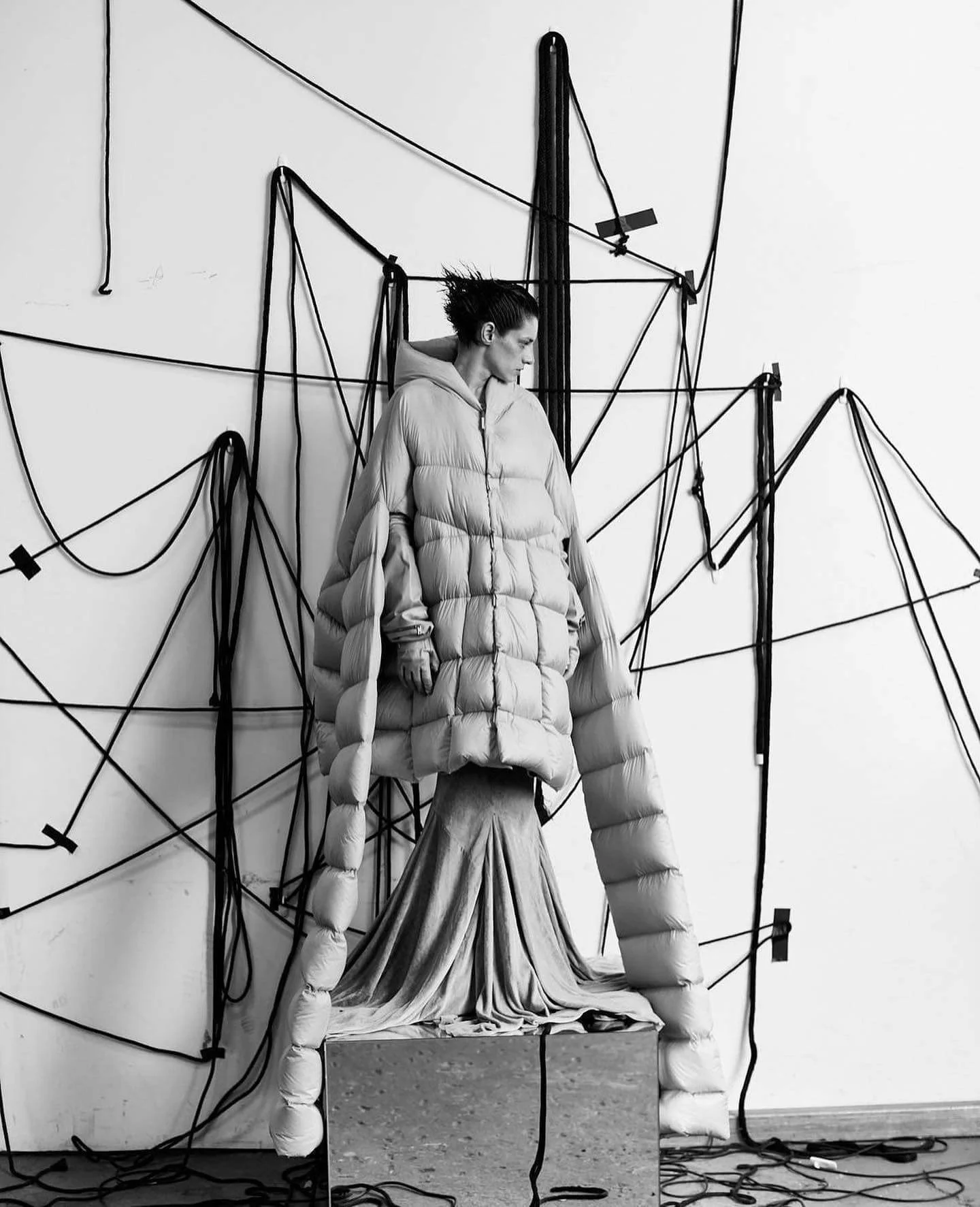 A black and white photo of a woman in a long puffy coat standing in front of a wall with interconnected black wires and some tape, with a draped fabric at her feet.