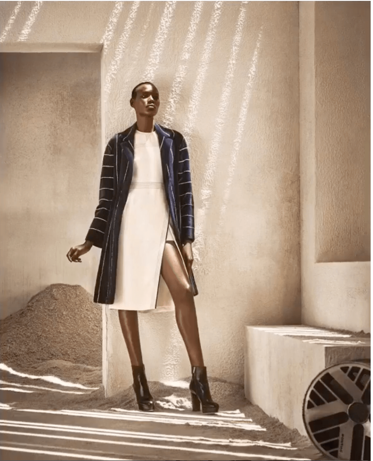 A woman in a white dress with a slit, black heeled boots, and a dark striped coat standing in a minimalist, textured room with shadows on the wall.