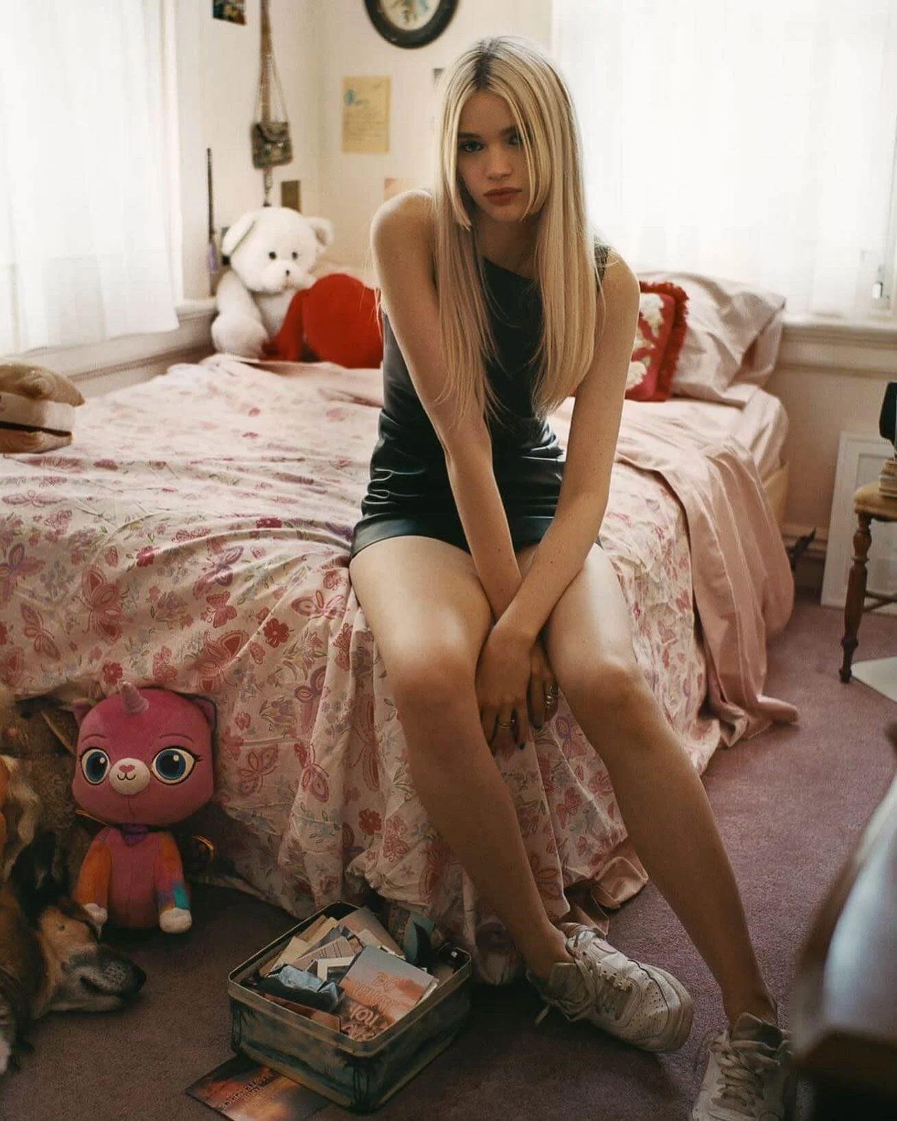 A young blonde woman is sitting on the edge of a bed in a bedroom, wearing a black sleeveless dress and white sneakers. The bed has a floral bedsheet and various stuffed animals, including a white teddy bear and a pink plush toy, are on and near the 