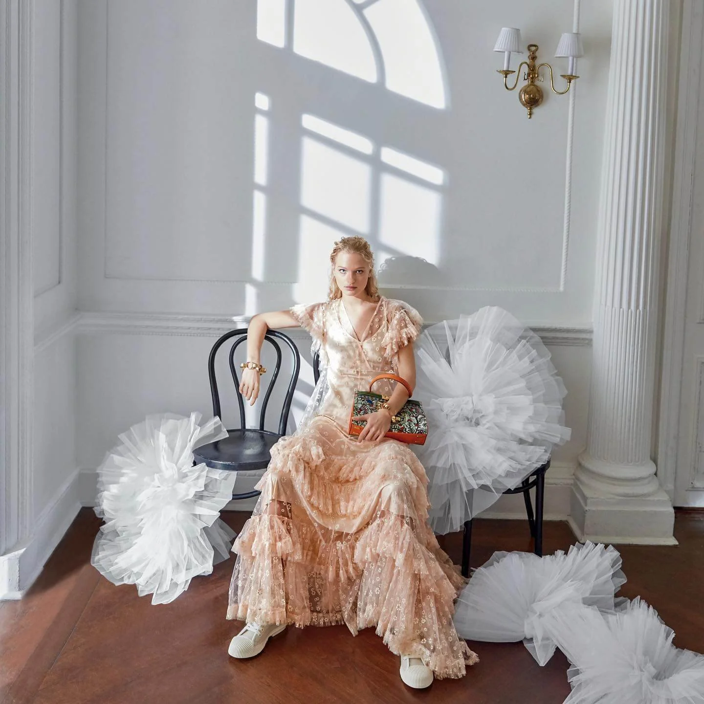 A young woman seated on a black chair in a white room with sunlight casting shadows through a window. She is wearing a peach-colored lace dress with ruffles, white sneakers, and accessorized with gold jewelry. She holds a colorful handbag in her lap 