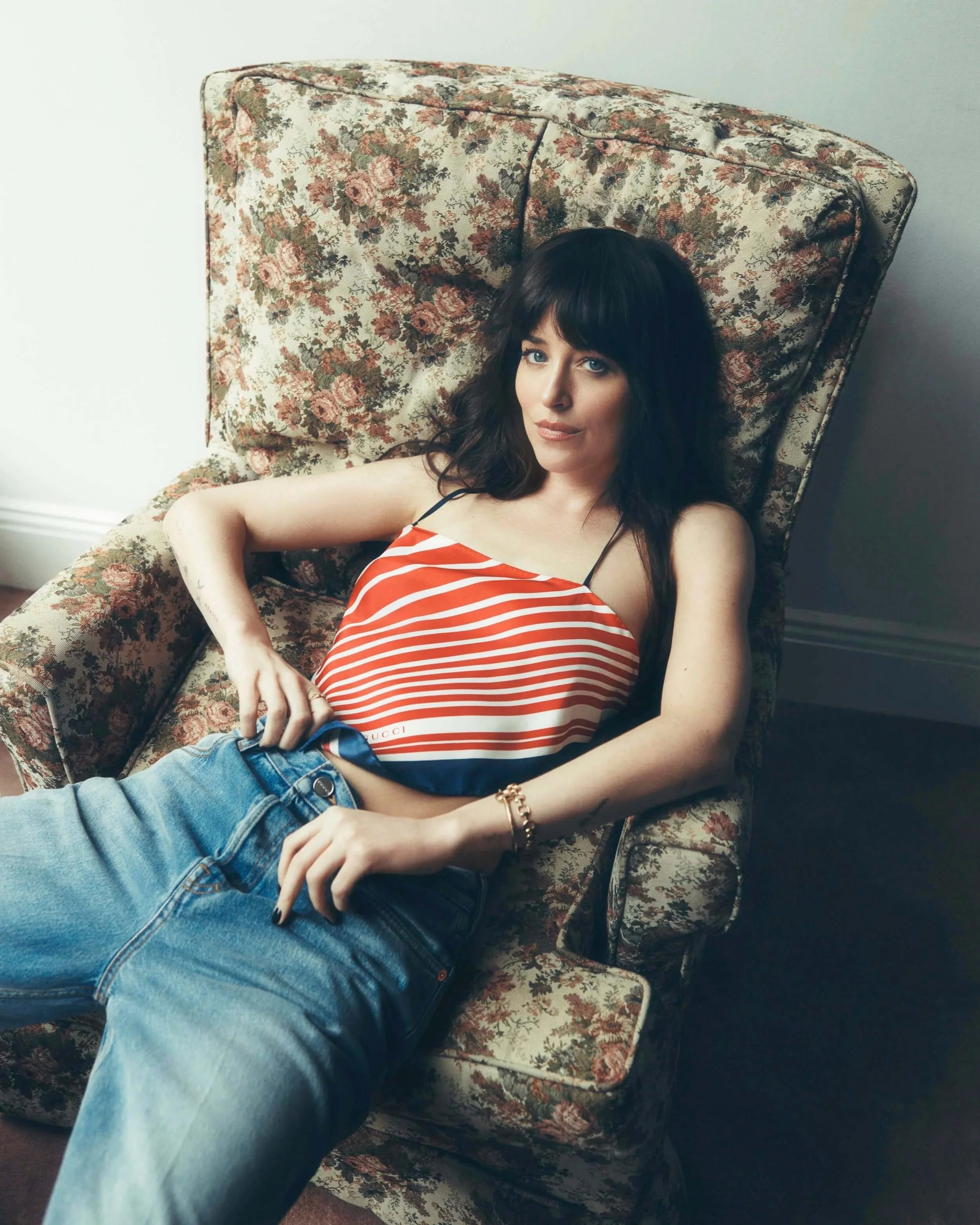 Woman with dark hair and bangs lying on a floral patterned vintage armchair, wearing a red and white striped top and blue jeans.