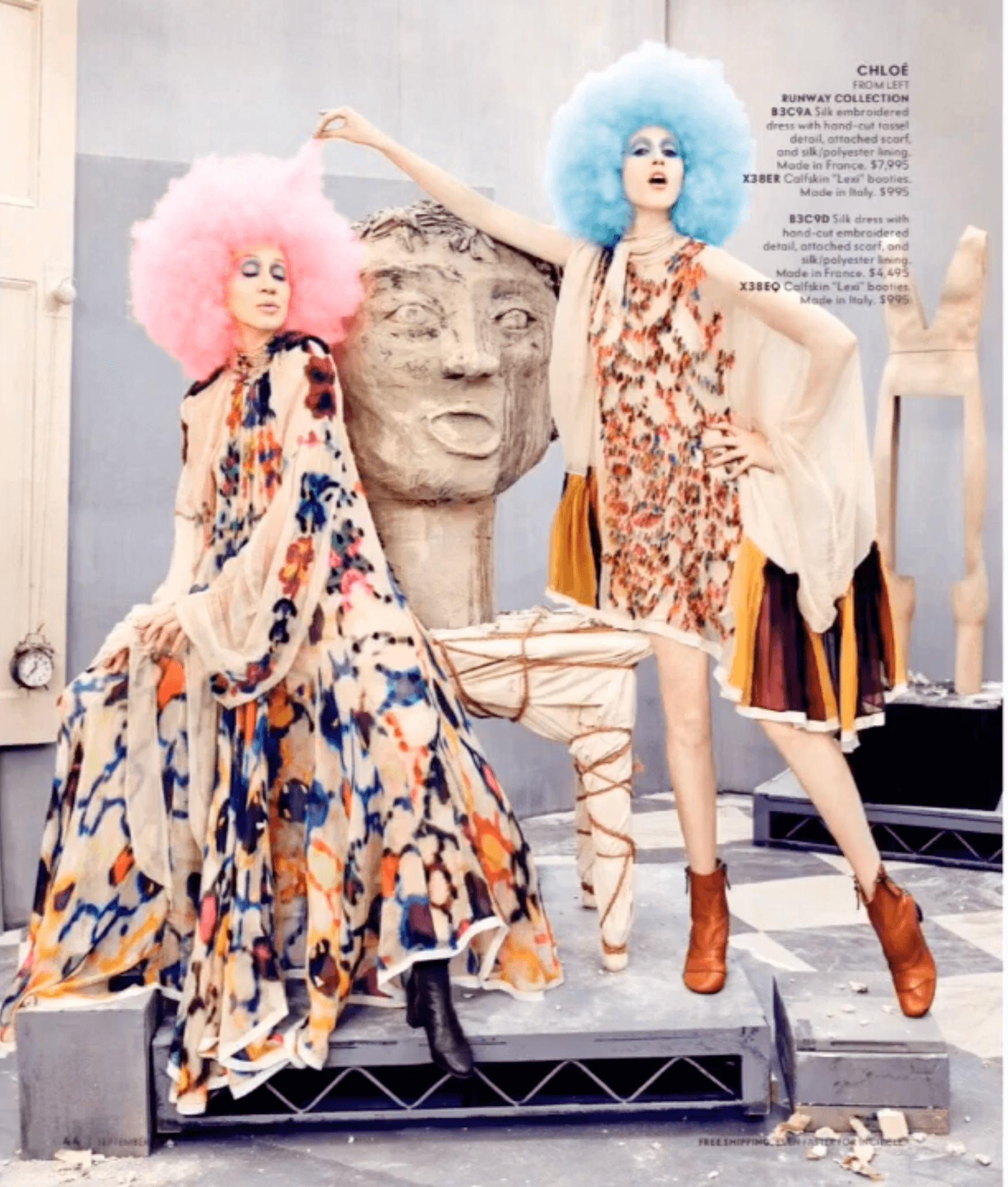 Two women with vibrant pink and blue afro wigs and colorful, patterned dresses standing next to a large sculpture of a human face in a studio setting.