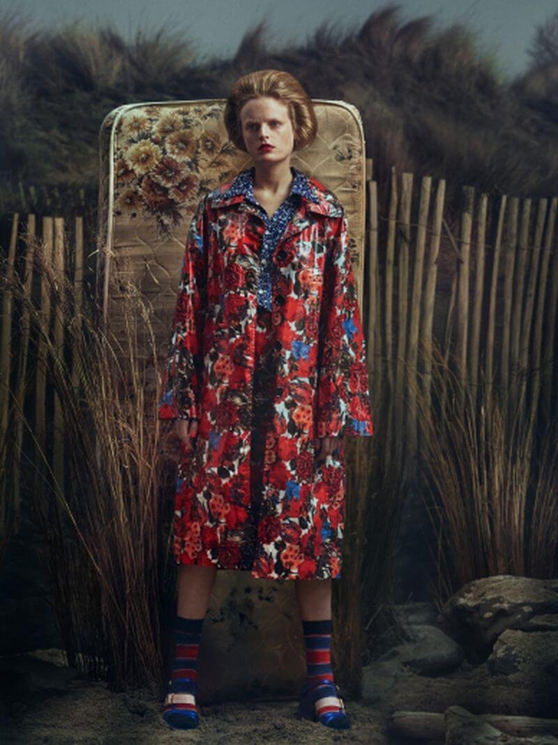 A woman with blond hair and red lipstick standing outdoors in front of a vintage mattress and wooden fence, wearing a long, colorful floral coat and striped socks.