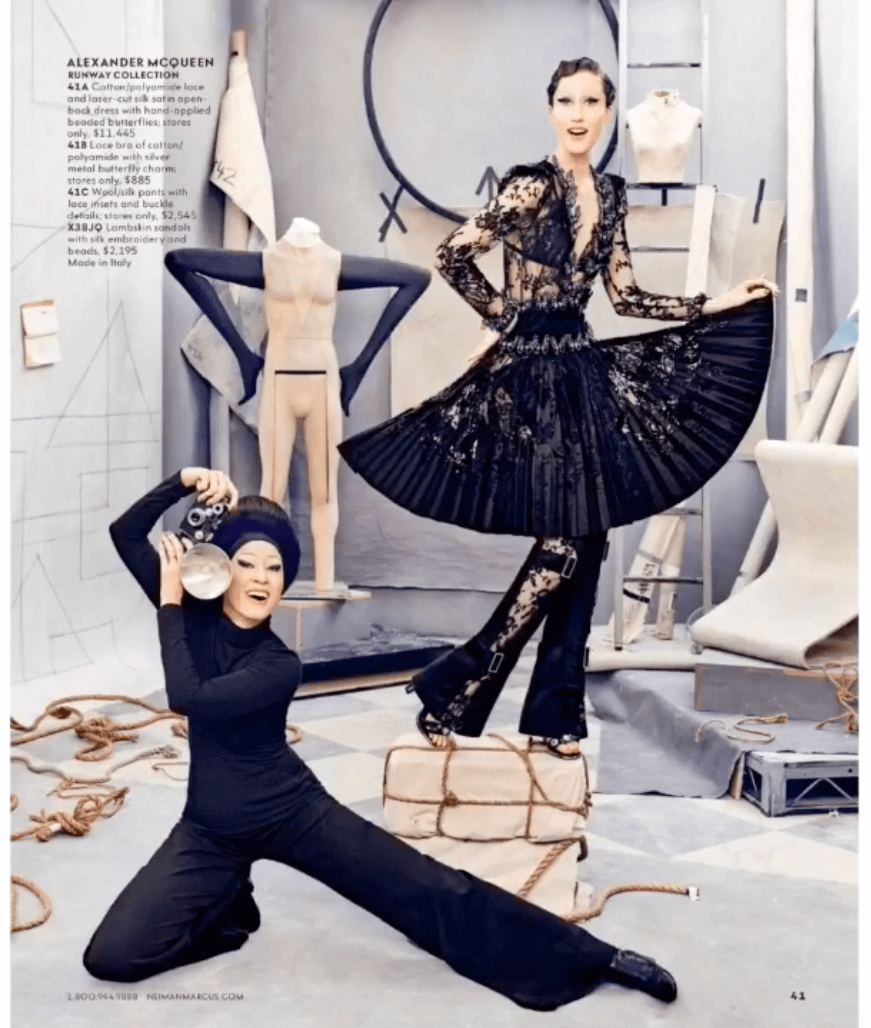 Model in a black lace dress with a pleated skirt performing on a platform, with a woman in black pants and top posing with a camera, in a studio with art supplies, ropes, and a mannequin.