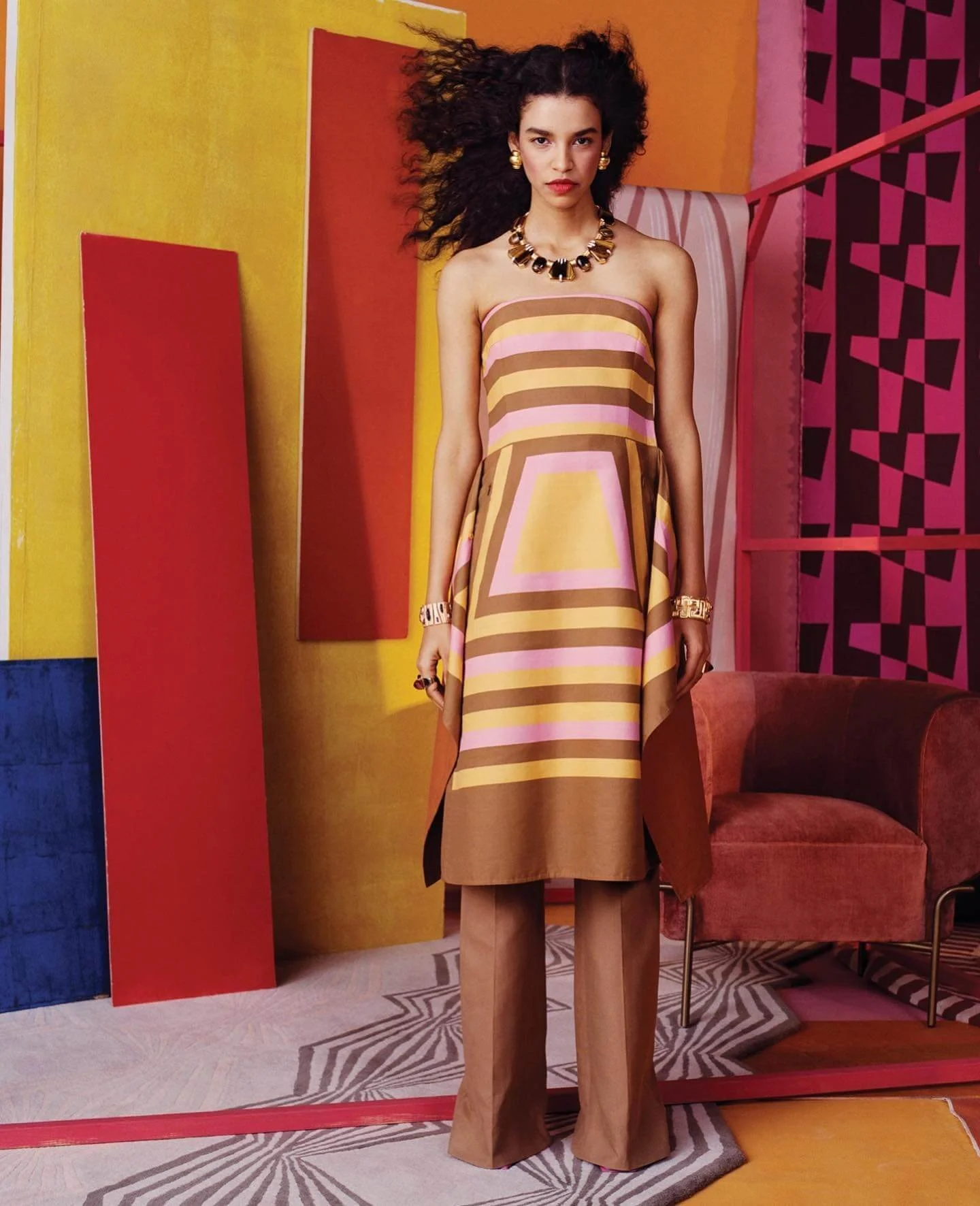 A woman standing in a colorful room wearing a strapless striped dress in shades of pink, yellow, and brown, with beige pants underneath, accessorized with gold jewelry including a necklace, earrings, bracelet, and rings, with an abstract geometric ba
