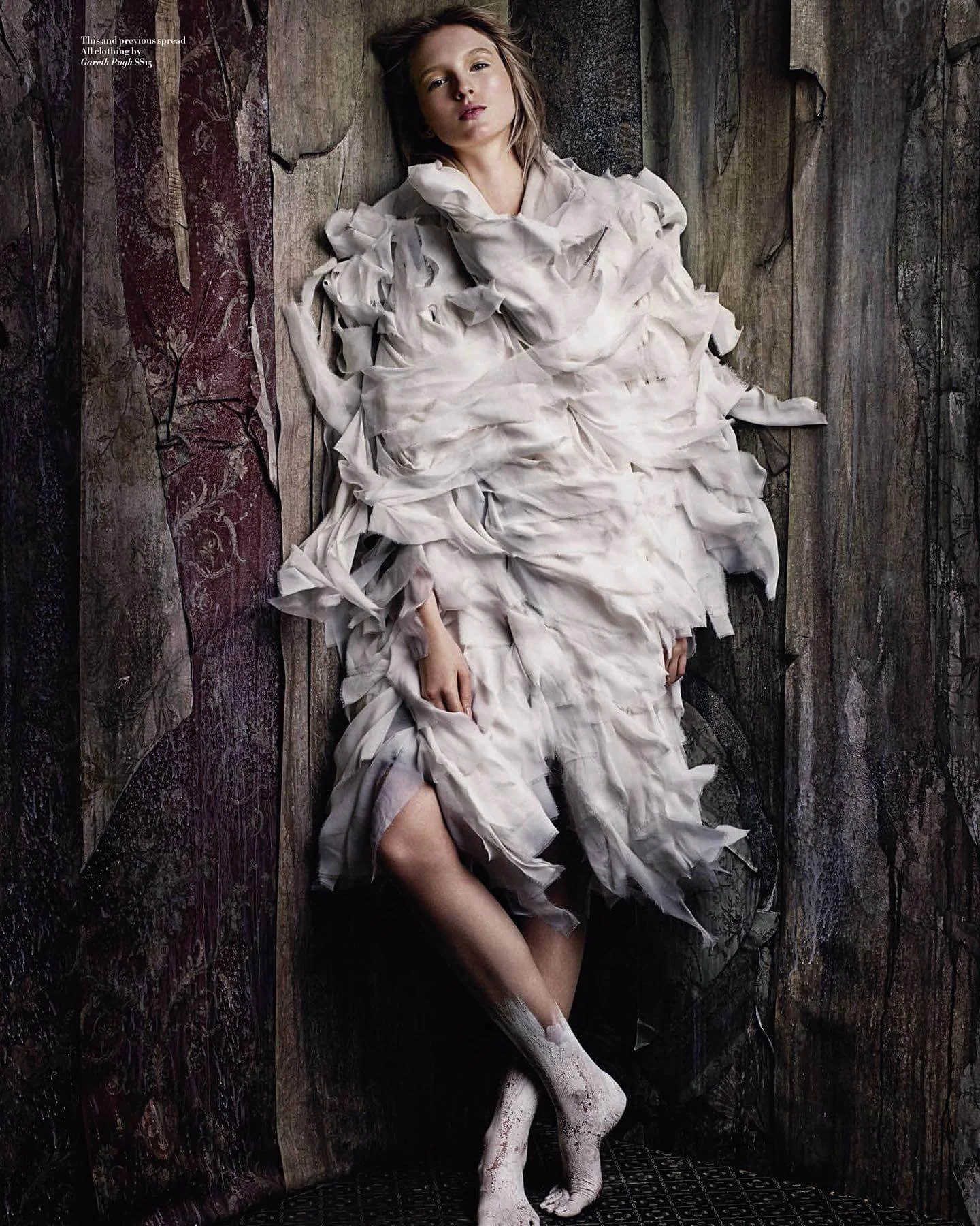 A young girl lying on the floor against a wooden wall, wearing a large, ruffled, white dress, with her head tilted slightly and one leg extended.
