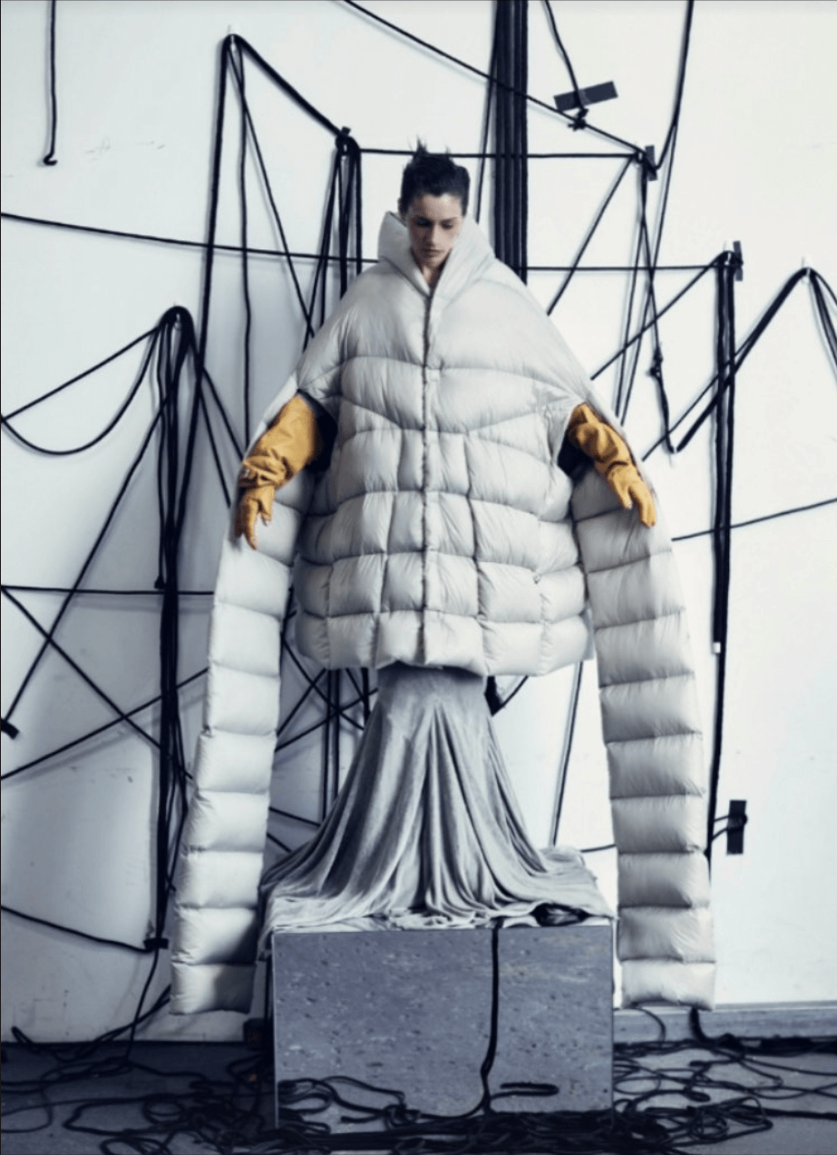 An art installation features a woman in a thick white puffer jacket with a high collar, wearing tan gloves. She stands in front of a white wall decorated with black wire sculptures resembling abstract geometric shapes. The woman appears to be emergin