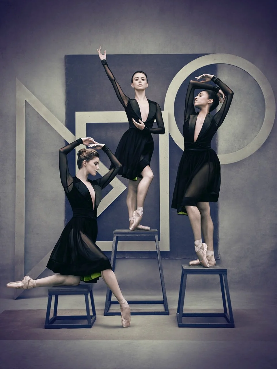 Three ballet dancers in black sheer dresses performing poses on stools and a step stool in a studio with geometric artwork in the background.