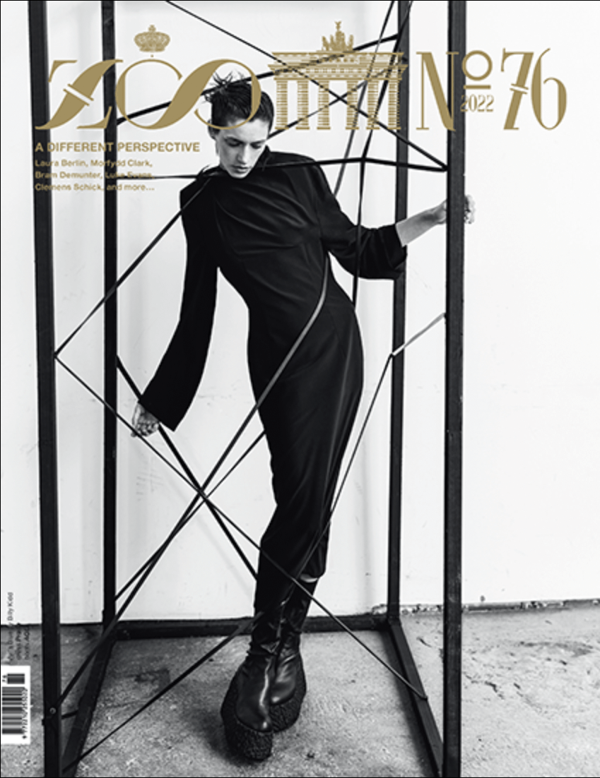 Black and white fashion magazine cover featuring a model in black clothing, standing inside a geometric metal frame against a white background, with gold text and logos at the top.