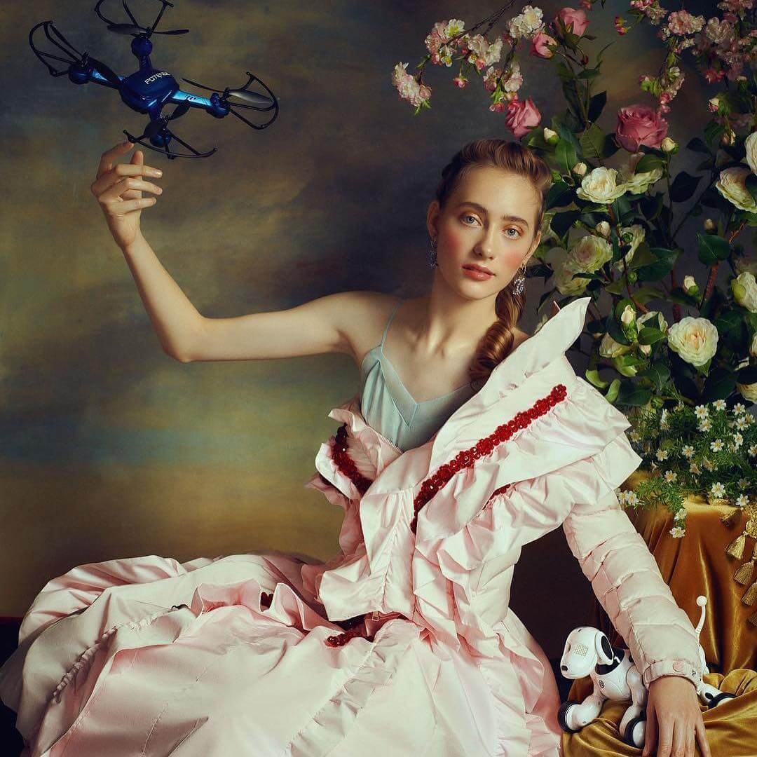 A woman in a pastel pink gown with ruffles and red embroidery, sitting next to a vase of white and pink flowers, holding a blue drone in one hand, with a small robotic dog on her lap.