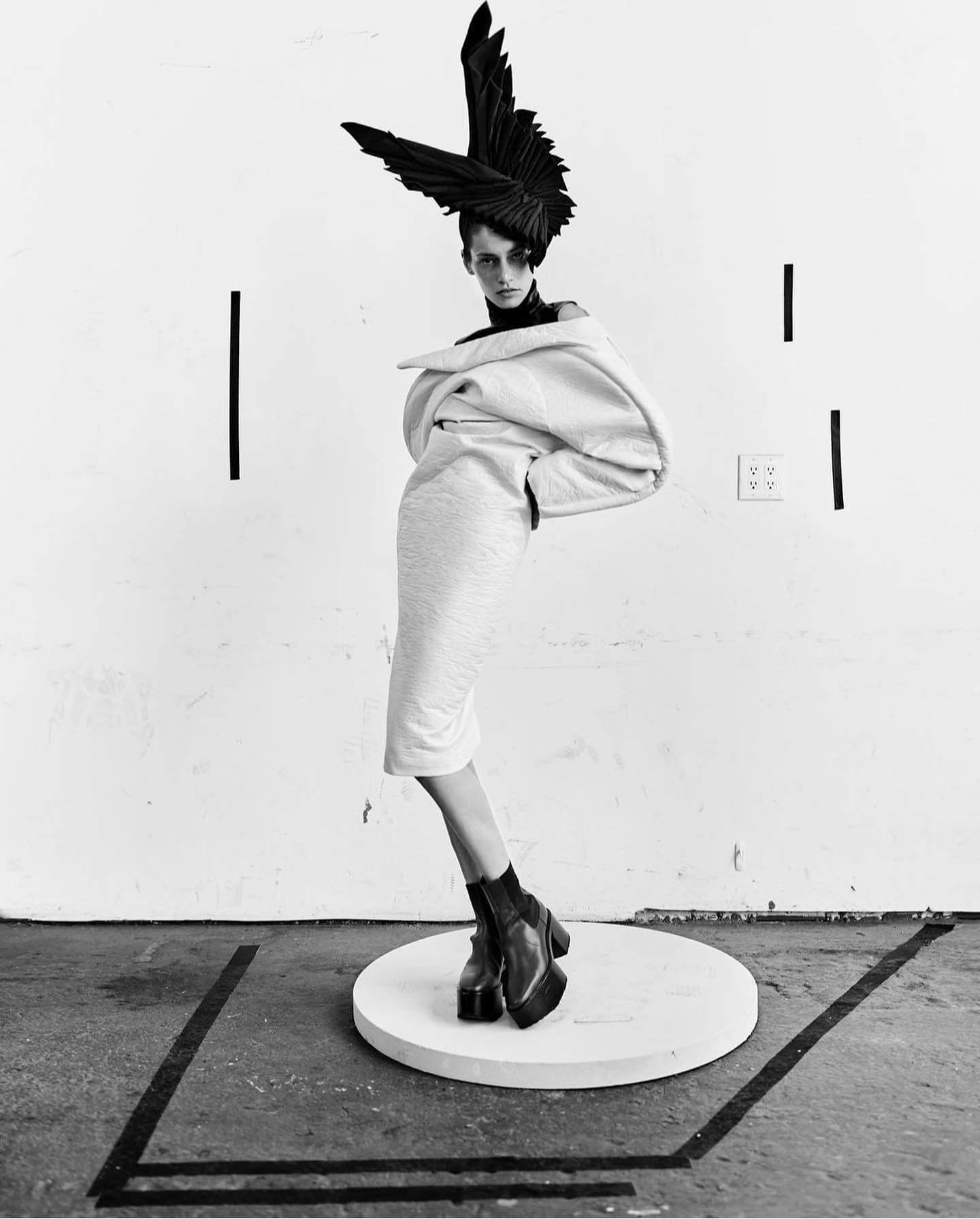 Black and white photo of a mannequin wearing chunky platform boots, a light-colored oversized hoodie, and a dark headpiece resembling large bird feathers, standing on a small round platform in front of a plain wall.