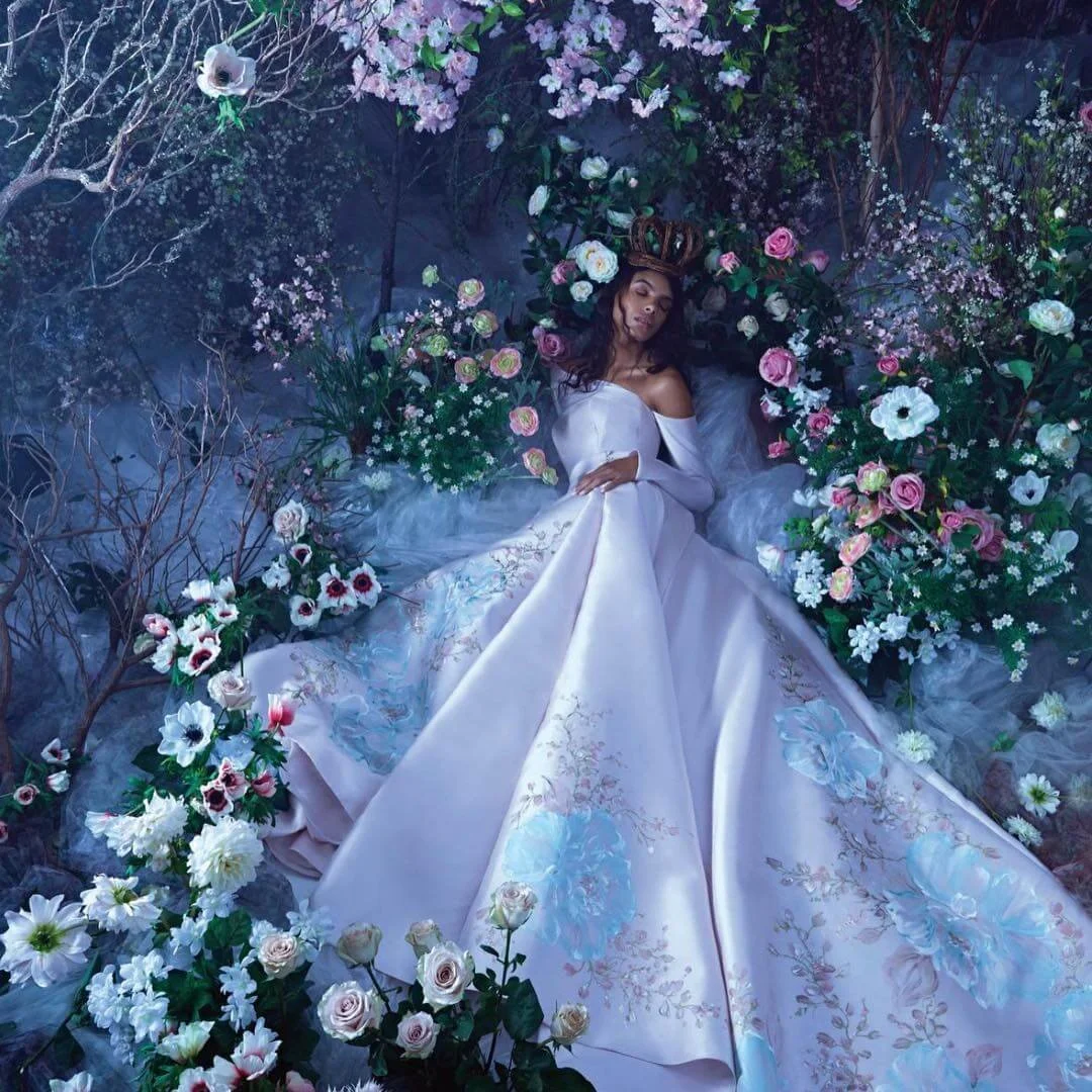 A woman in a white wedding gown lying on the ground surrounded by pink and white flowers and greenery, with her eyes closed and a crown on her head.