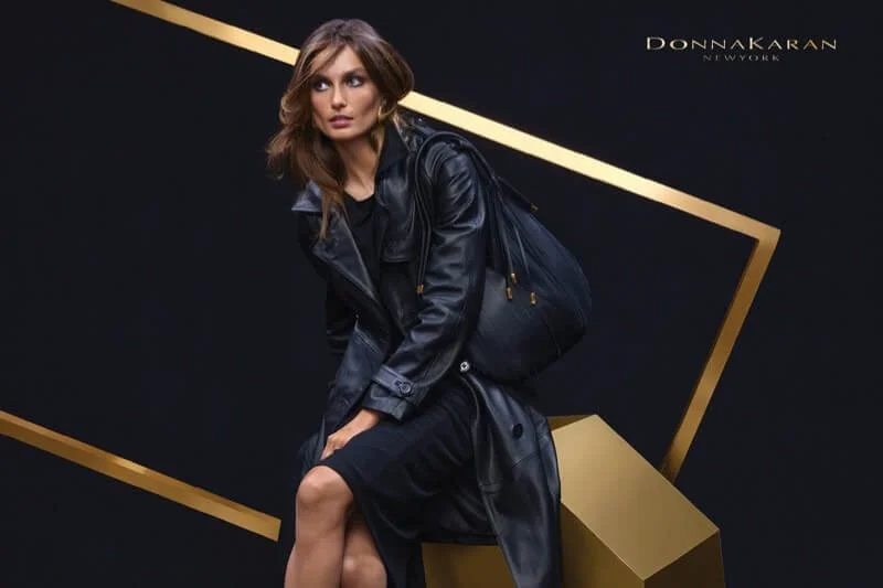 A woman with brown hair wearing a black leather jacket and black dress, sitting on a gold geometric block against a black background, with "Donnakaran Network" written in the upper right corner.