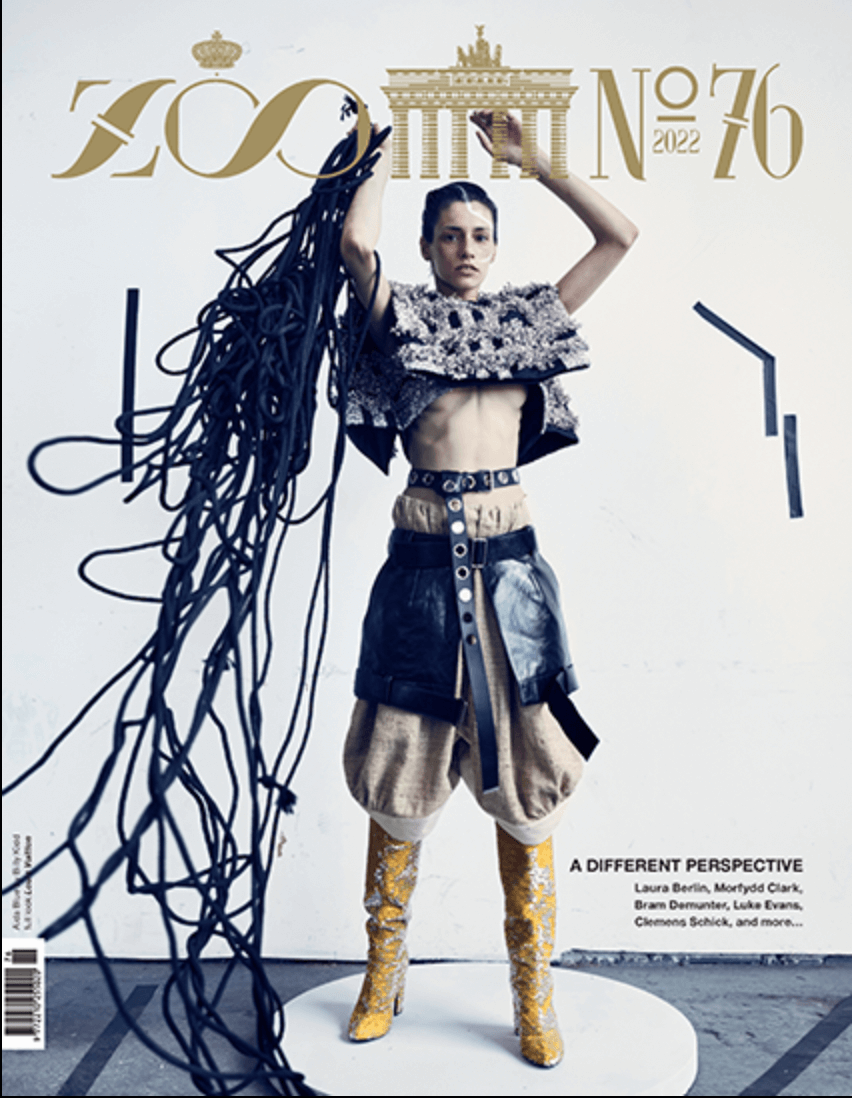 Fashion model in eclectic clothing standing against a white wall with artwork, including a graphic of the Brandenburg Gate and text for a fashion magazine cover, with long black cords hanging from her raised arm.