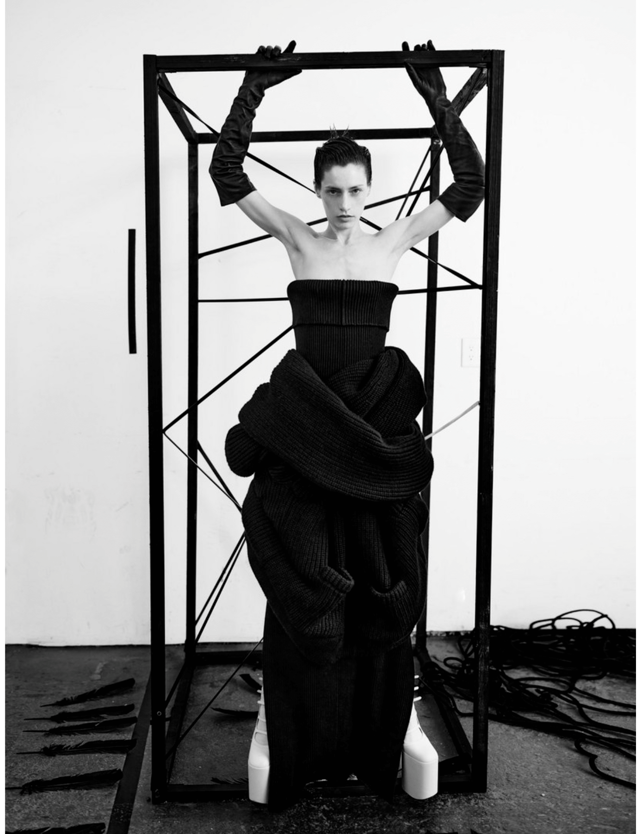 A black and white photograph of a woman with short hair, wearing a strapless top and pants made of large knitted fabric, standing inside a geometric metal frame. She is holding the top of the frame with both hands and wearing long gloves.