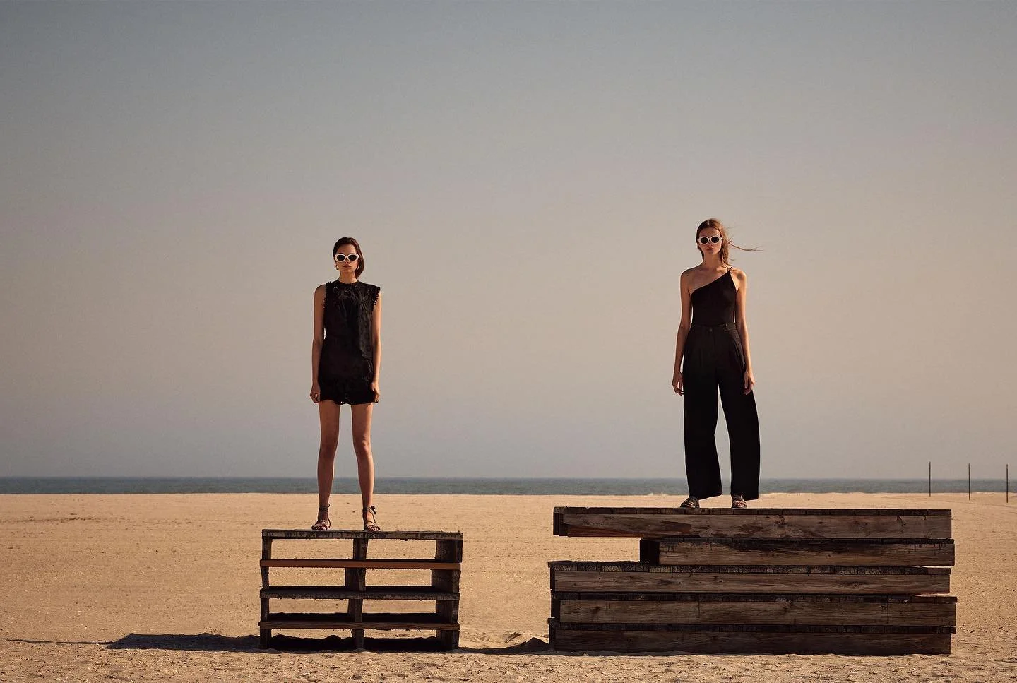 Two women standing on wooden pallets on a sandy beach with the ocean and sky in the background, both wearing black outfits and sunglasses.
