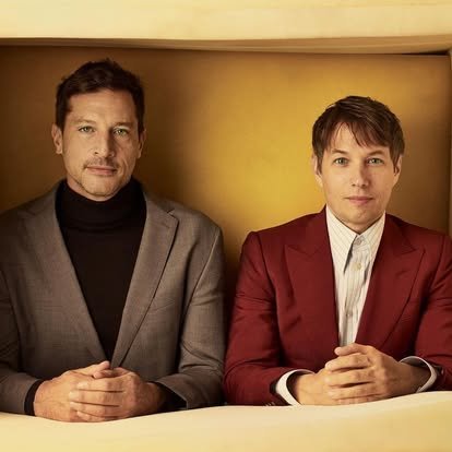 Two men sitting at a table with their hands clasped in front of them, wearing suits and posing for a photo.