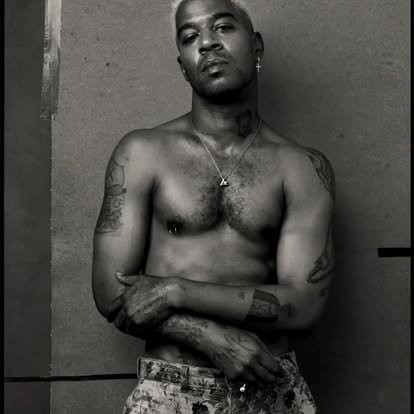 A shirtless man with tattoos on his arms, wearing a necklace and earring, standing with arms crossed against a plain wall in black and white.